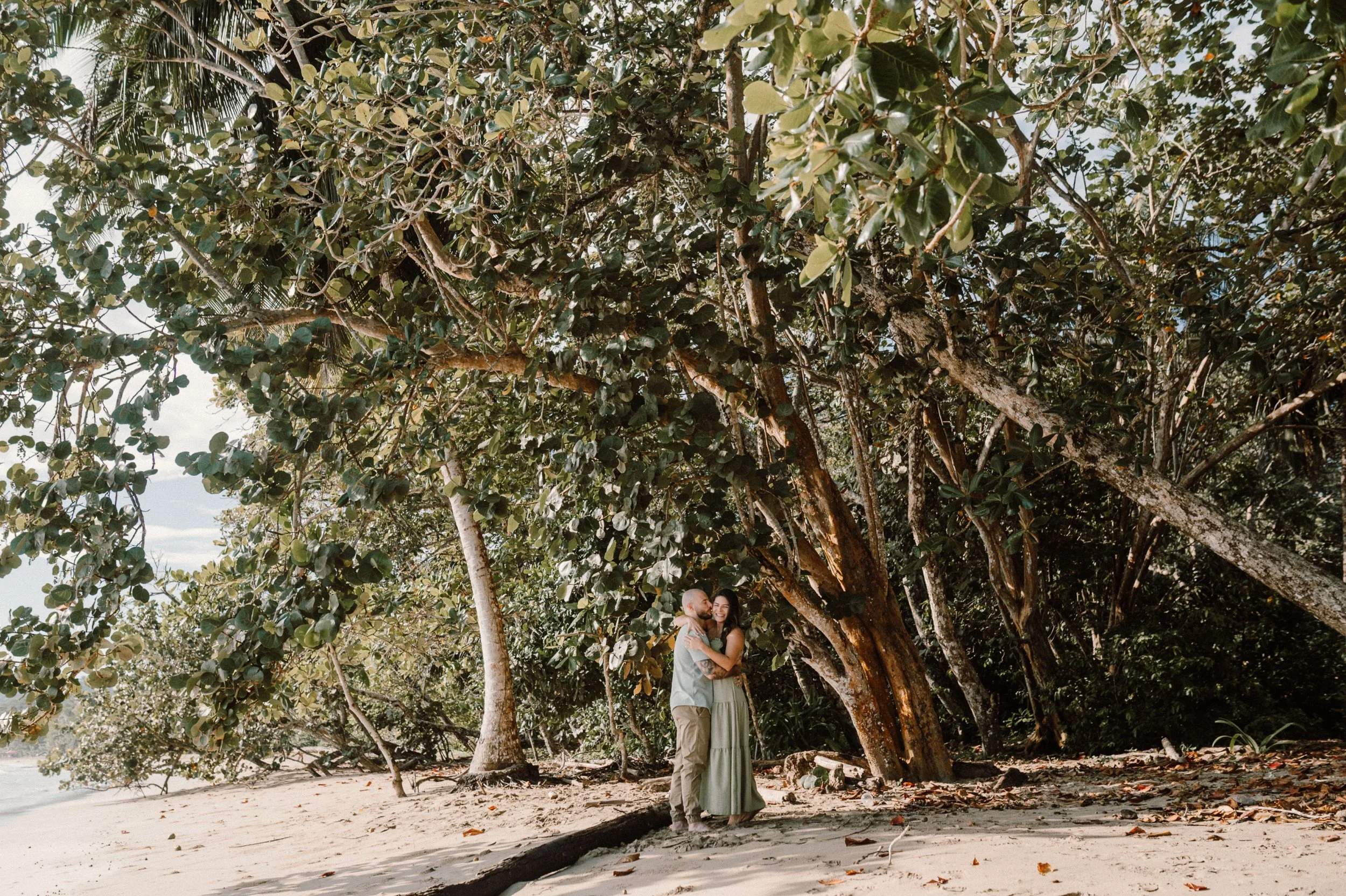 Una pareja abrazada en la playa junto a árboles grandes y frondosos bajo un cielo despejado.
