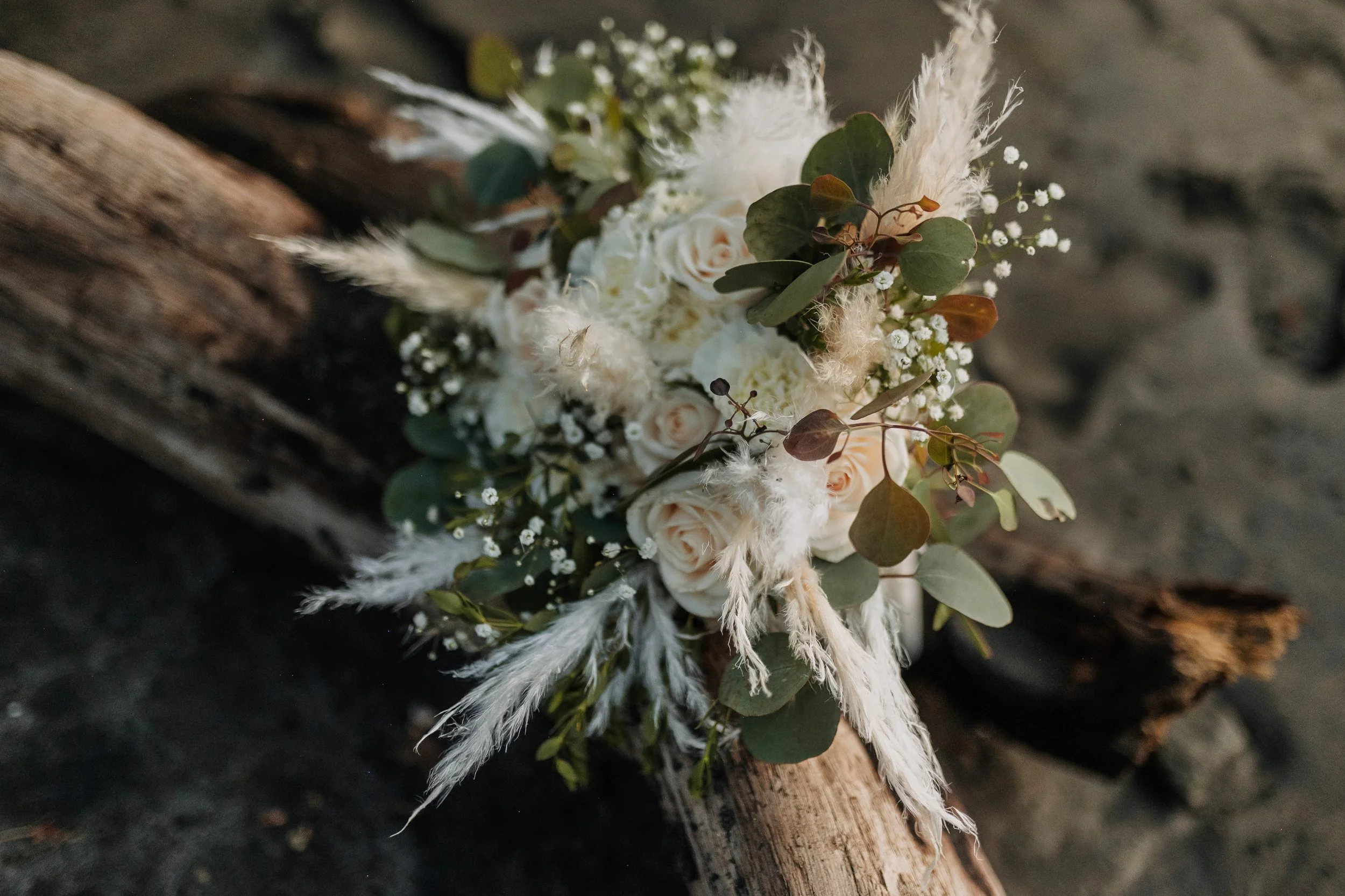 Ramo de flores blancas con hojas verdes y plumas blancas, colocado sobre un tronco de árbol en la playa.
