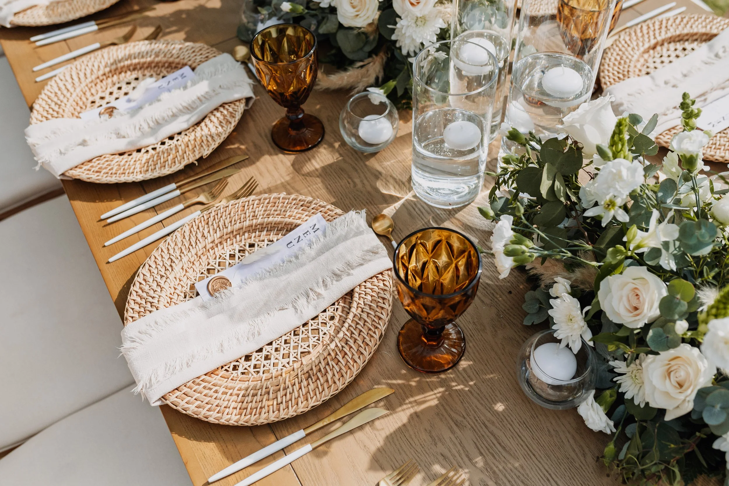 Mesa decorada con platos de ratán, cubiertos dorados, copas de vino ámbar, vasos de agua con velas flotando, flores blancas y verdes, y servilletas blancas con flecos, lista para una comida especial.