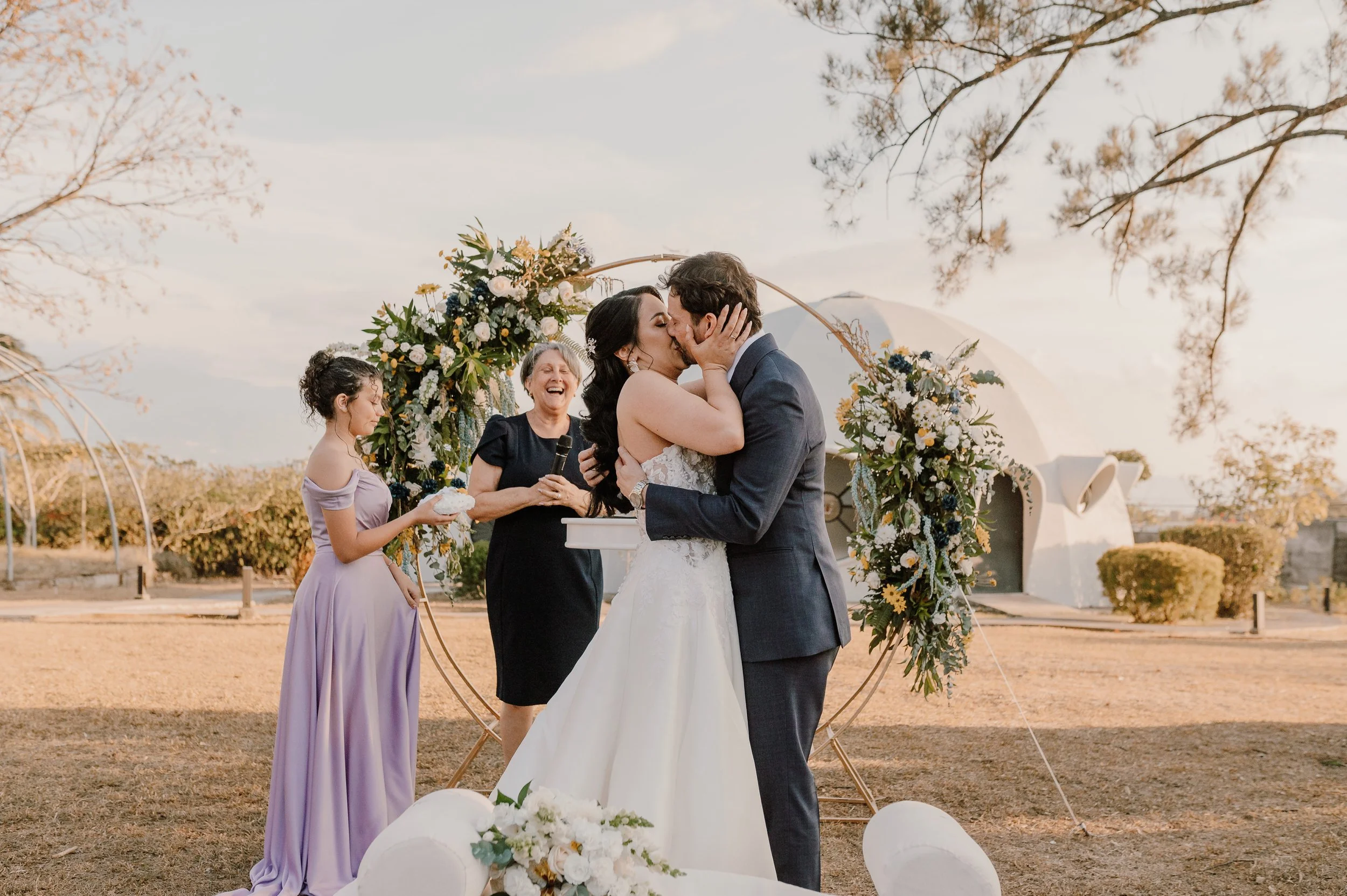 Pareja de recién casados besándose en una ceremonia al aire libre con decoraciones florales y un arco, mientras una mujer joven y una mujer mayor los observan, en un entorno natural con árboles y un edificio blanco en el fondo.