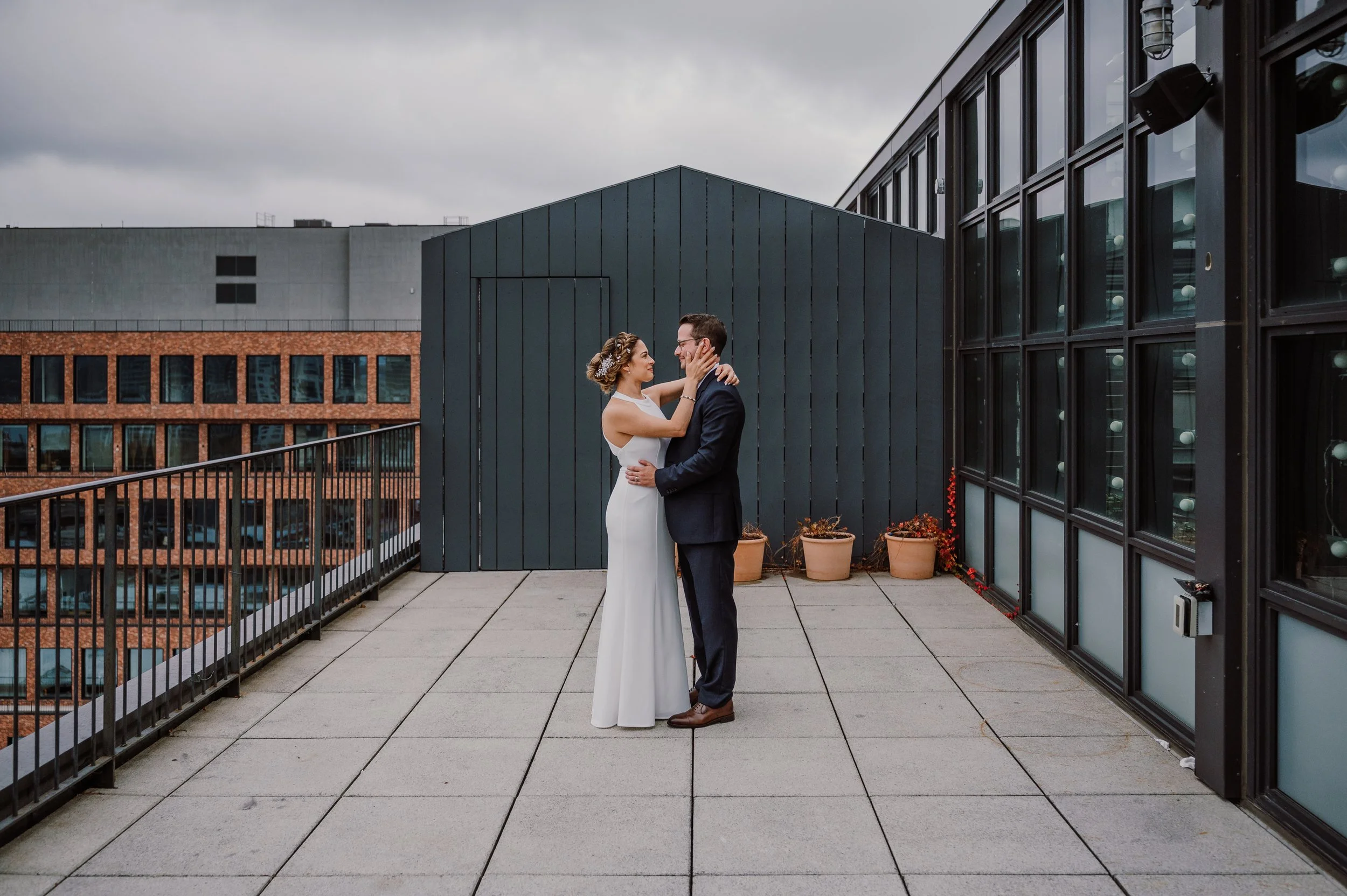 Pareja de novios en la azotea, ella con vestido blanco y él con traje oscuro, en un día nublado, en un entorno urbano con edificios de fondo y plantas en macetas.