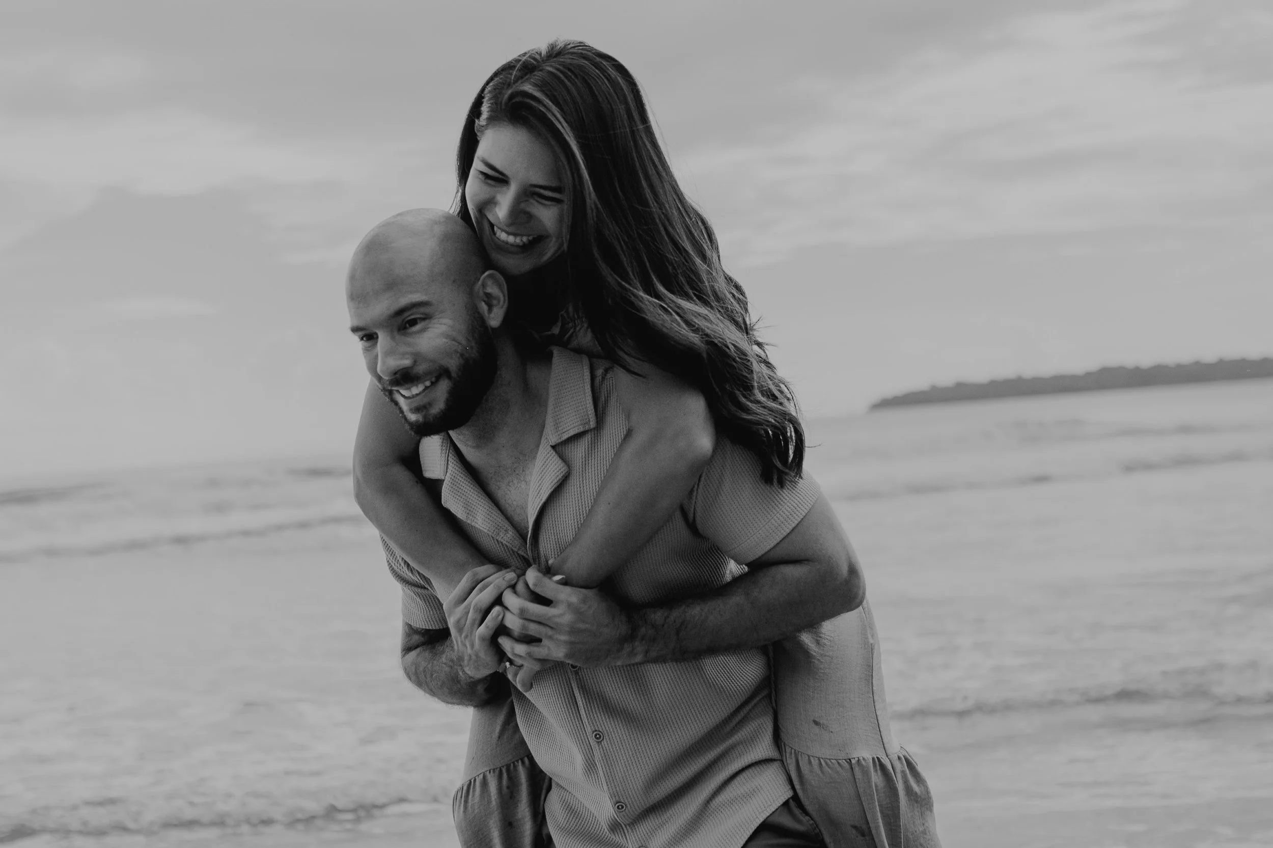 Pareja sonriendo en la playa, el hombre lleva a la mujer en la espalda, vista en blanco y negro.