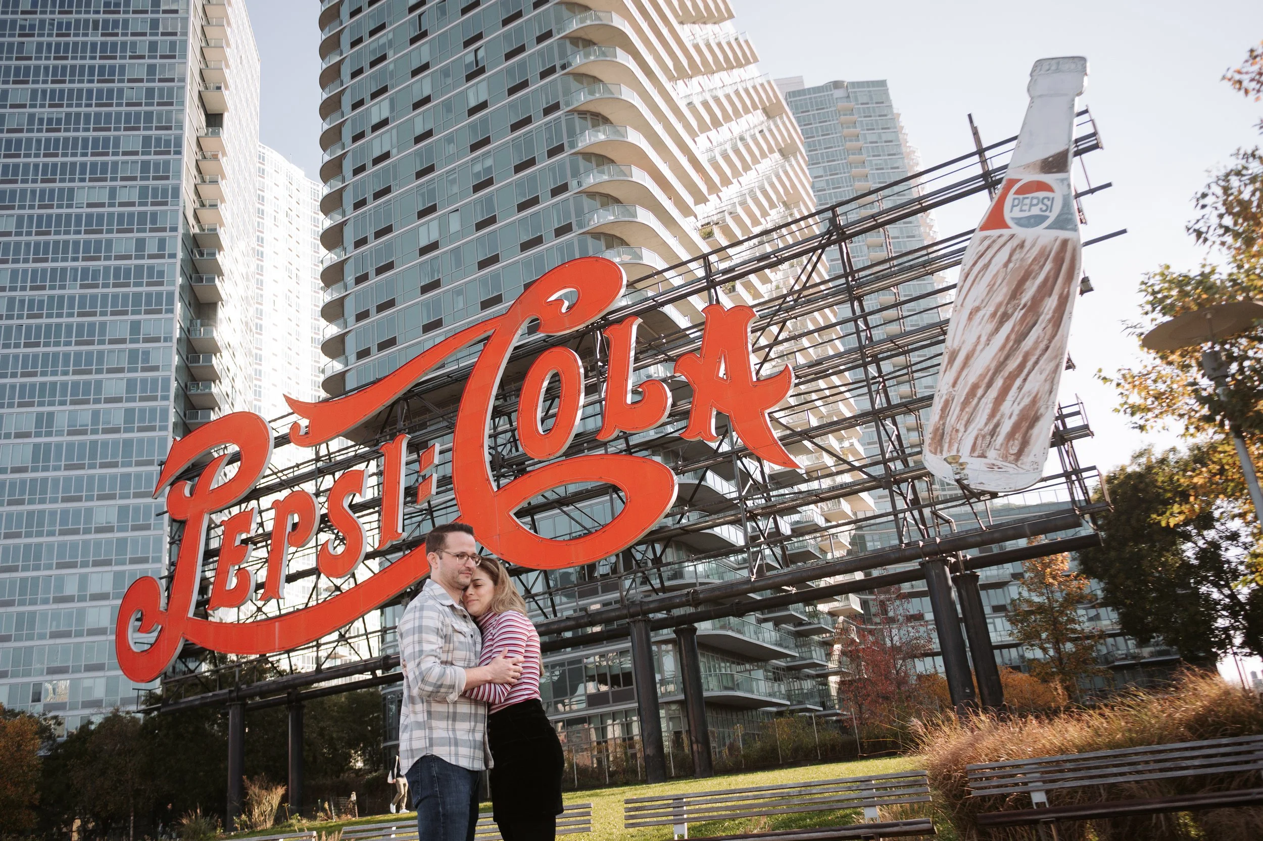 Una pareja abrazada frente a un cartel grande de Pepsi Cola en un entorno urbano con edificios modernos de apartamentos y árboles.