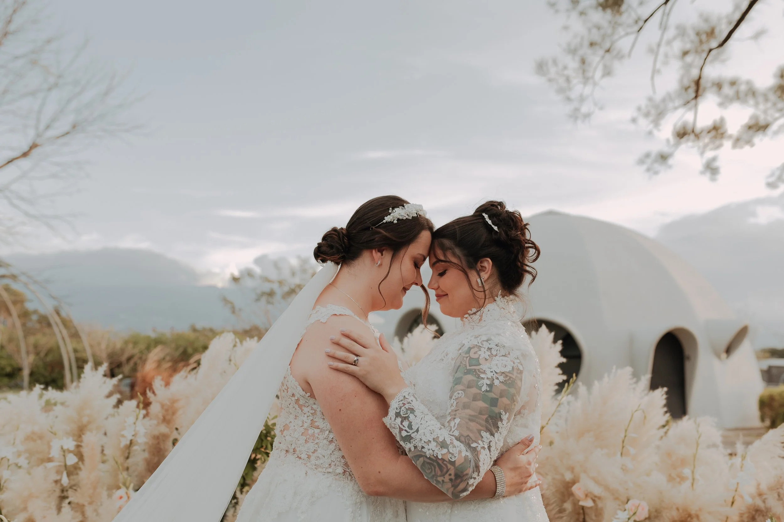 Dos mujeres en un abrazo, vestidas con trajes de novia, en un entorno al aire libre con plantas y un edificio blanco futurista al fondo.