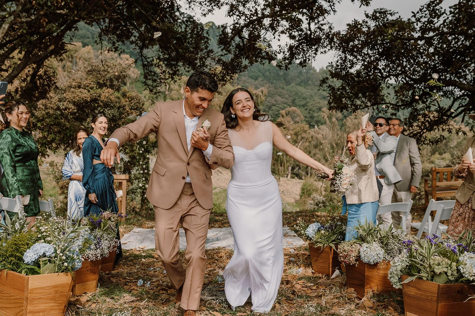 Una pareja de novios caminando juntos en una ceremonia al aire libre, rodeados de amigos y familiares, con árboles y montañas de fondo.
