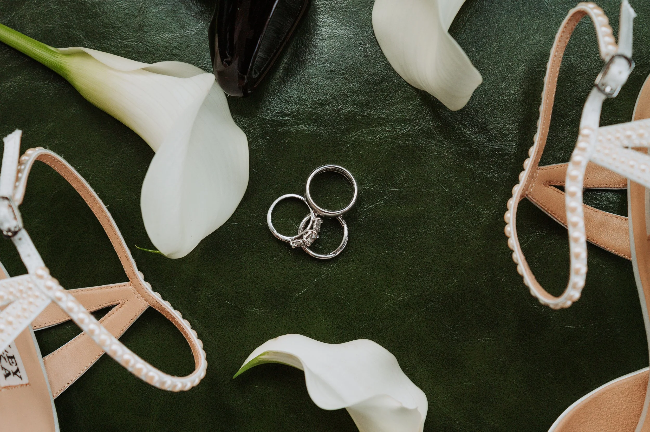Zapatos de tacón beige, dos anillos plateados, calla lilies blancas, fondo oscuro.