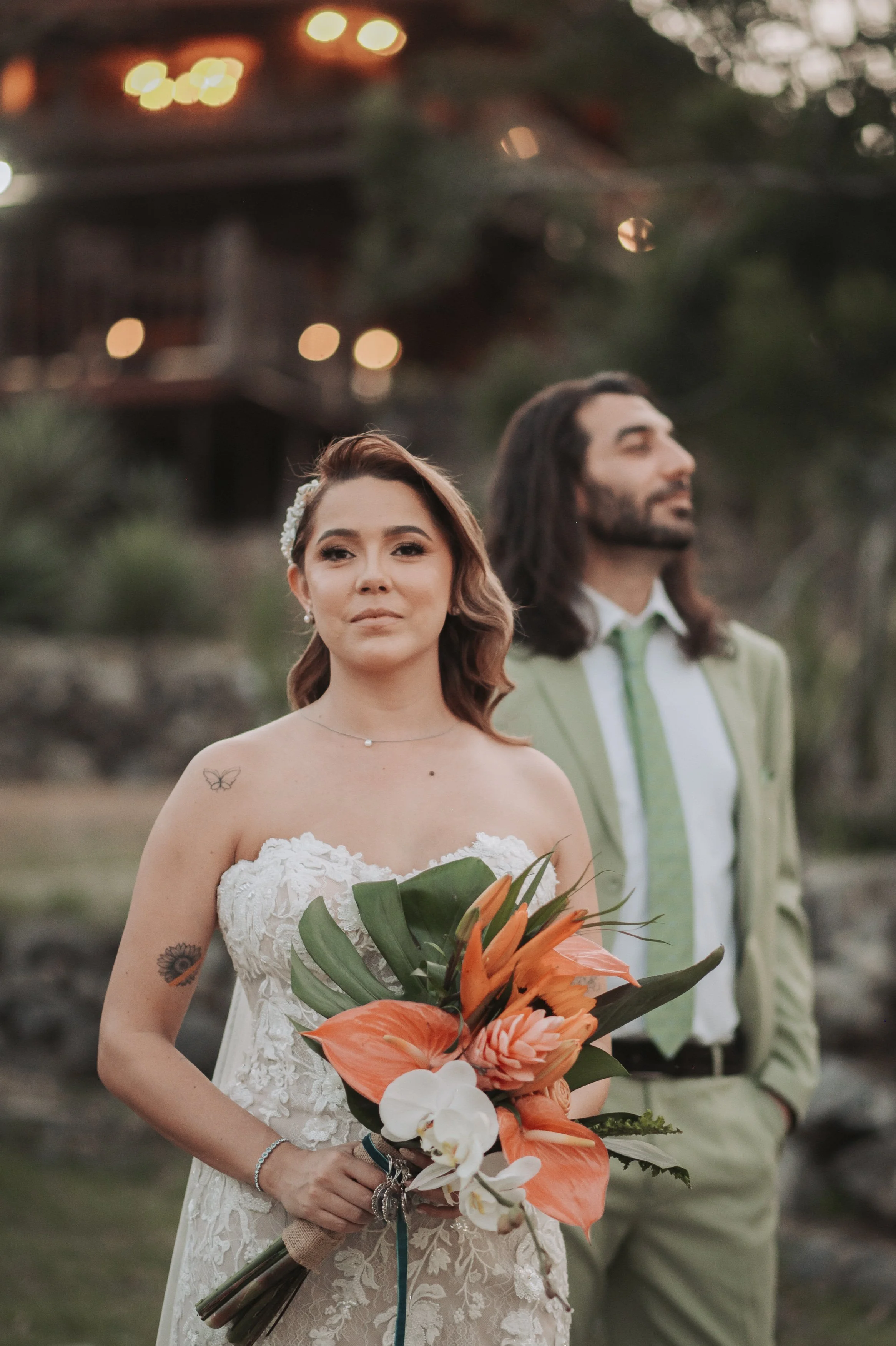 Mujer en vestido de novia sosteniendo un ramo de flores naranjas y blancas, con un hombre de fondo con chaqueta verde claro y corbata, en un entorno natural.