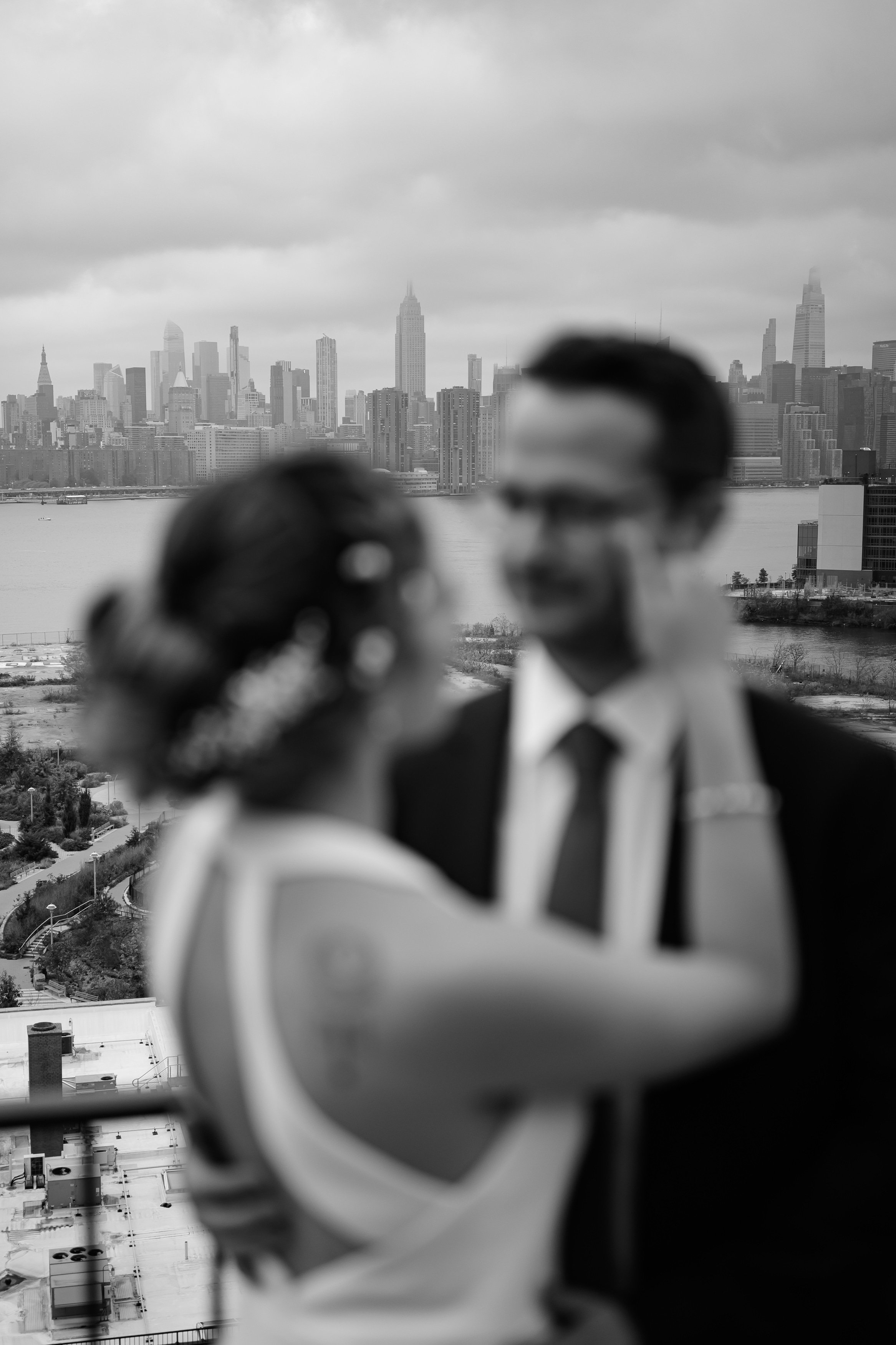 Una pareja de boda en primer plano, desenfocados, con el skyline de la ciudad de Nueva York al fondo, en un día nublado.