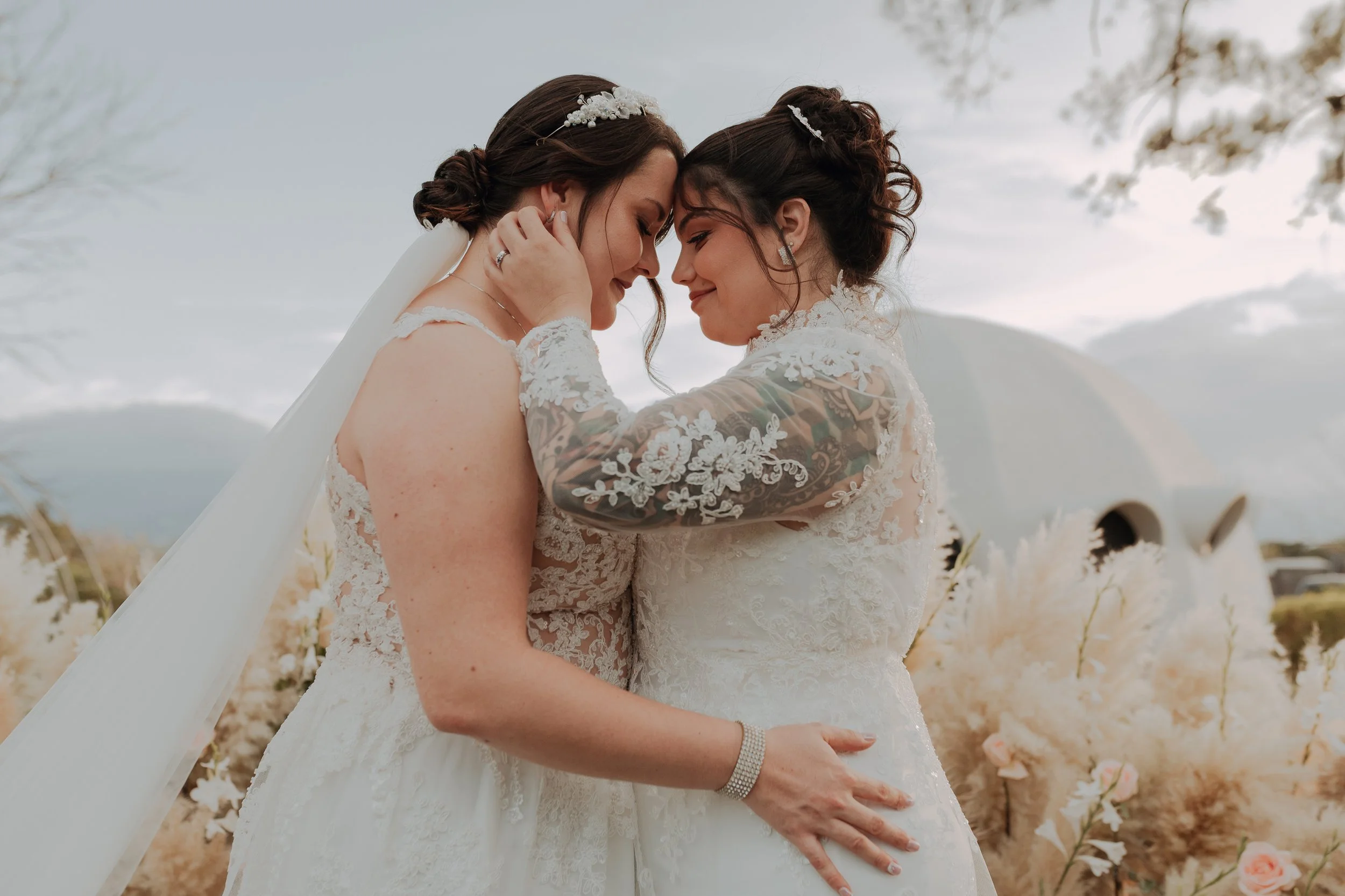 Dos novias con vestidos de boda en un abrazo, tocándose la frente, en un entorno natural con flores y árboles.