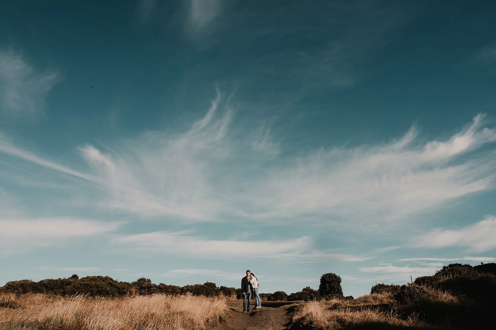 Una pareja caminando en un sendero en un campo abierto con arbustos y árboles dispersos bajo un cielo azul con nubes delgadas.