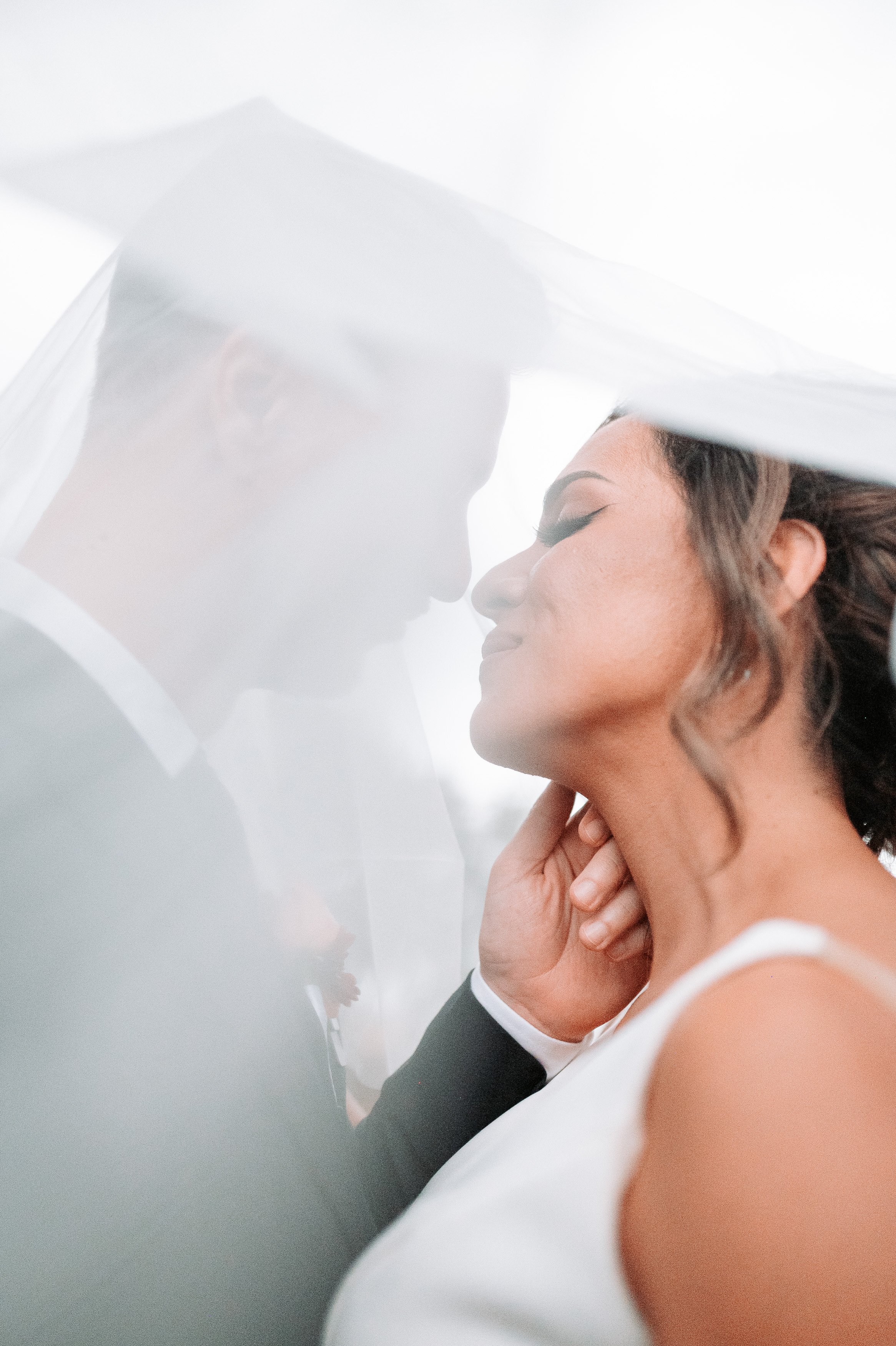 Una pareja de recién casados bajo un velo de novia, en una pose romántica con la frente tocándose, con ojos cerrados y sonriendo suavemente.