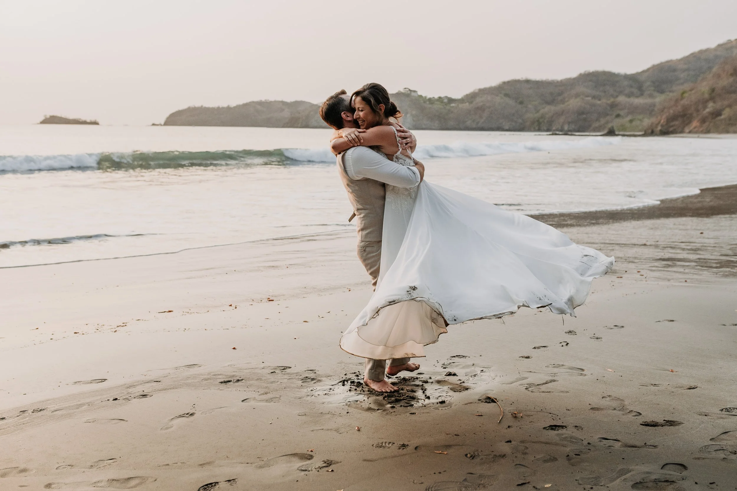 Pareja de recién casados en la playa, el hombre levantando a la mujer en una escena romántica al atardecer.