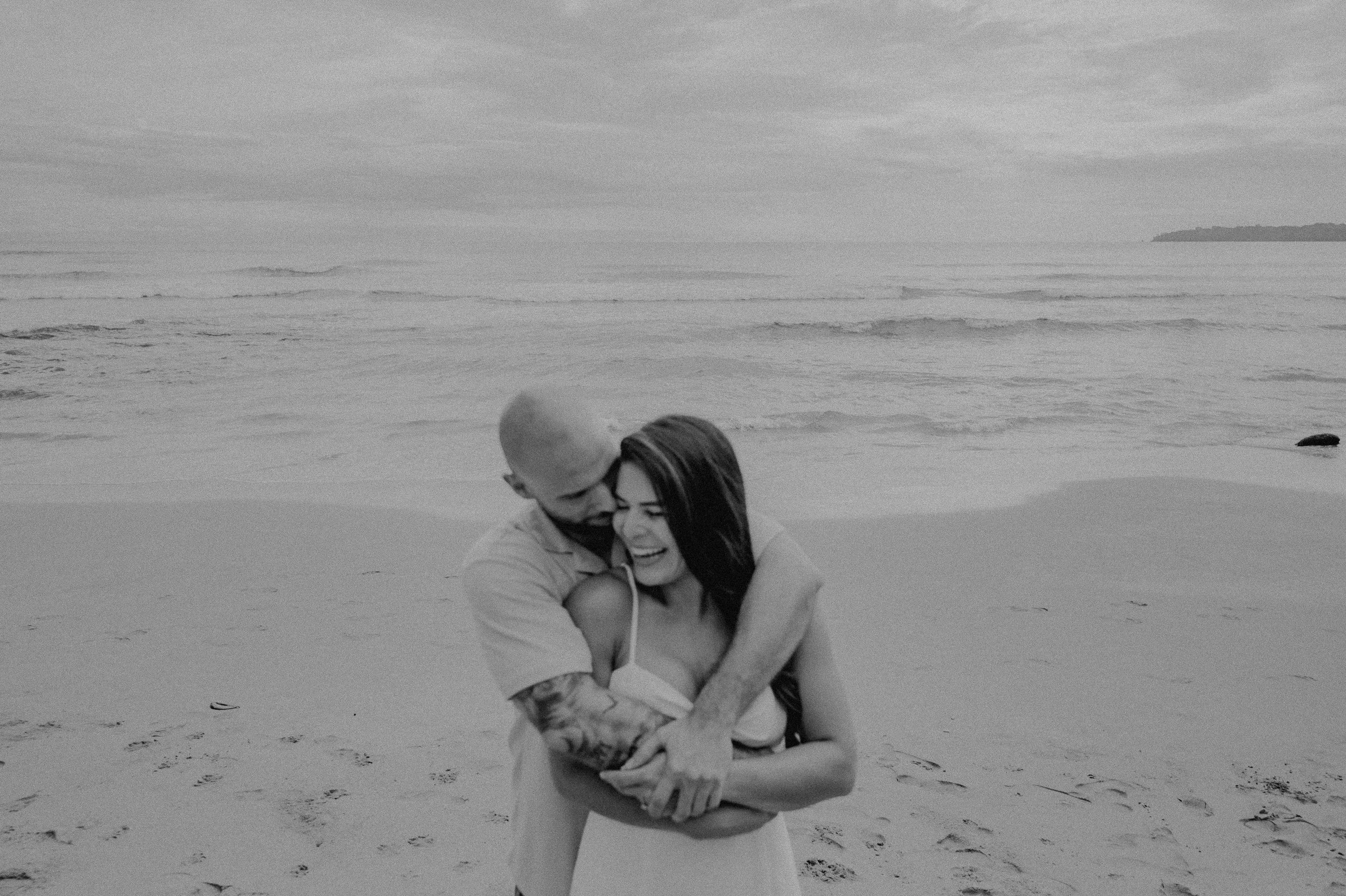 Una pareja feliz abrazándose en la playa, con el mar y el cielo en el fondo.