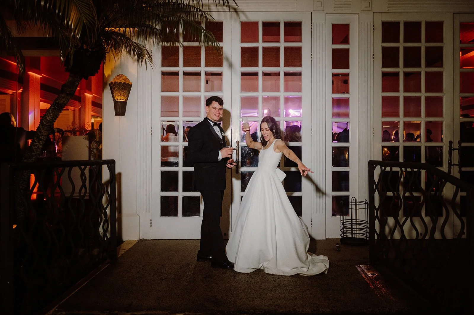 Pareja en una celebración, la mujer con vestido de novia y el hombre con traje, ambos sonriendo y brindando con copas, en un lugar decorado con luz de neón rosada y ambiente festivo.