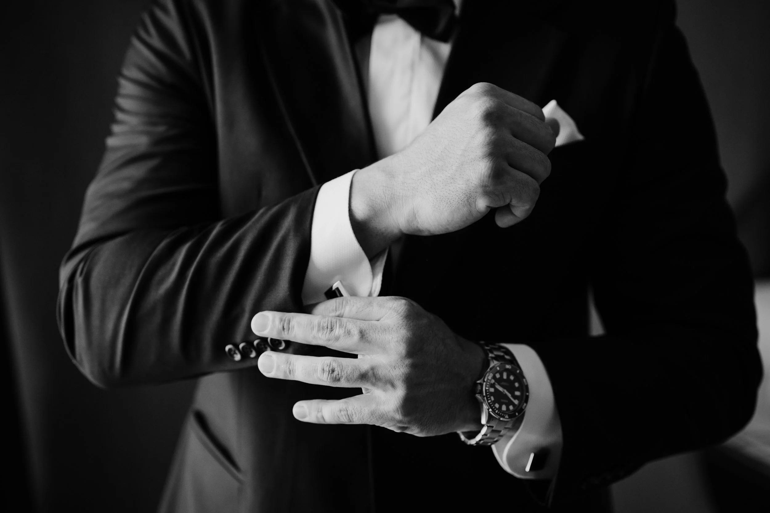 Un hombre de traje ajustándose la forma de la ropa, con un reloj en la muñeca, en una pose elegante en un fondo oscuro.