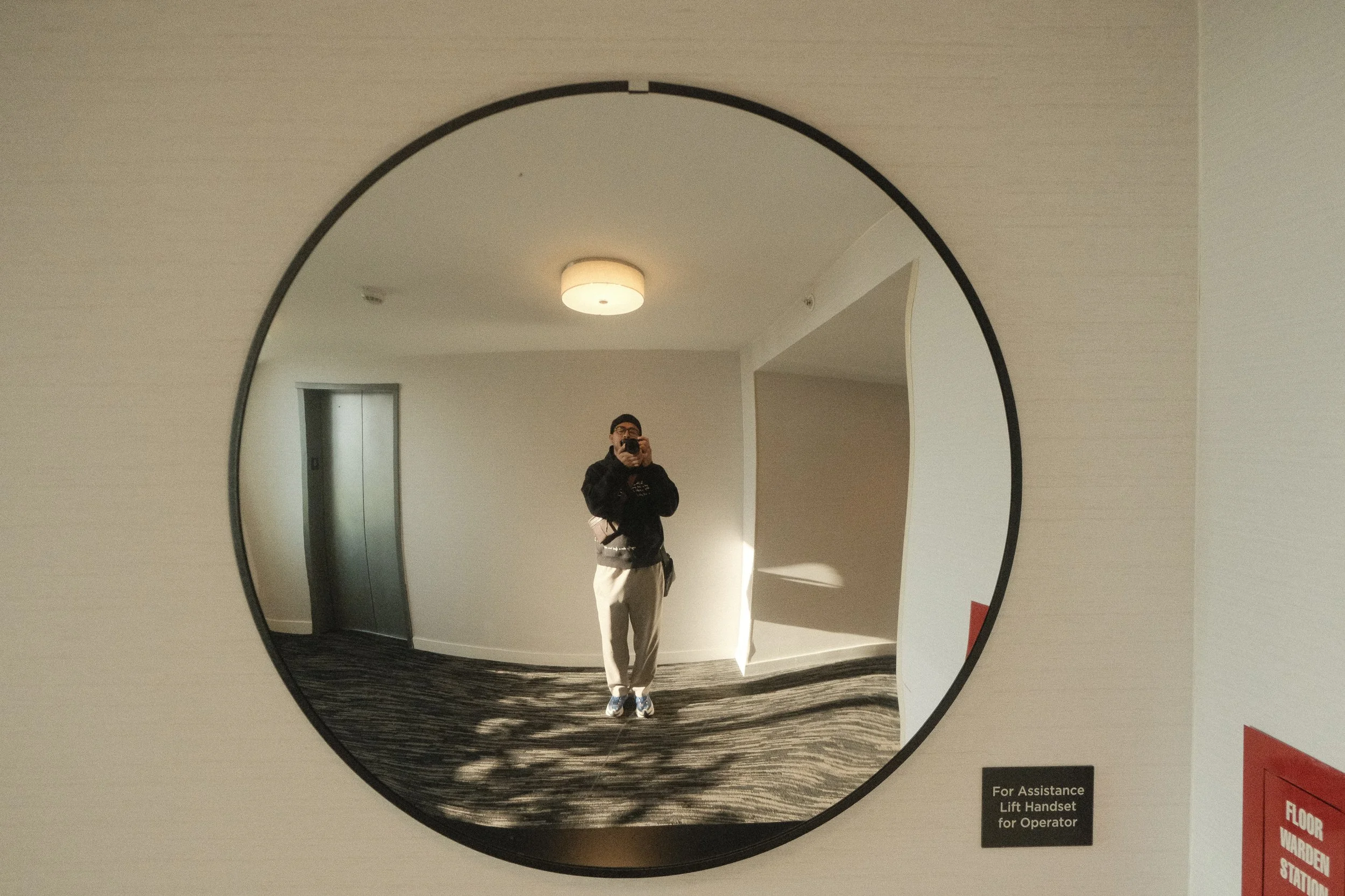 Espejo redondo en la pared que refleja a una persona con gafas, camisa negra y pantalones claros, tomando una selfie con teléfono móvil, en un ambiente interno con techo iluminado y puertas de ascensor.