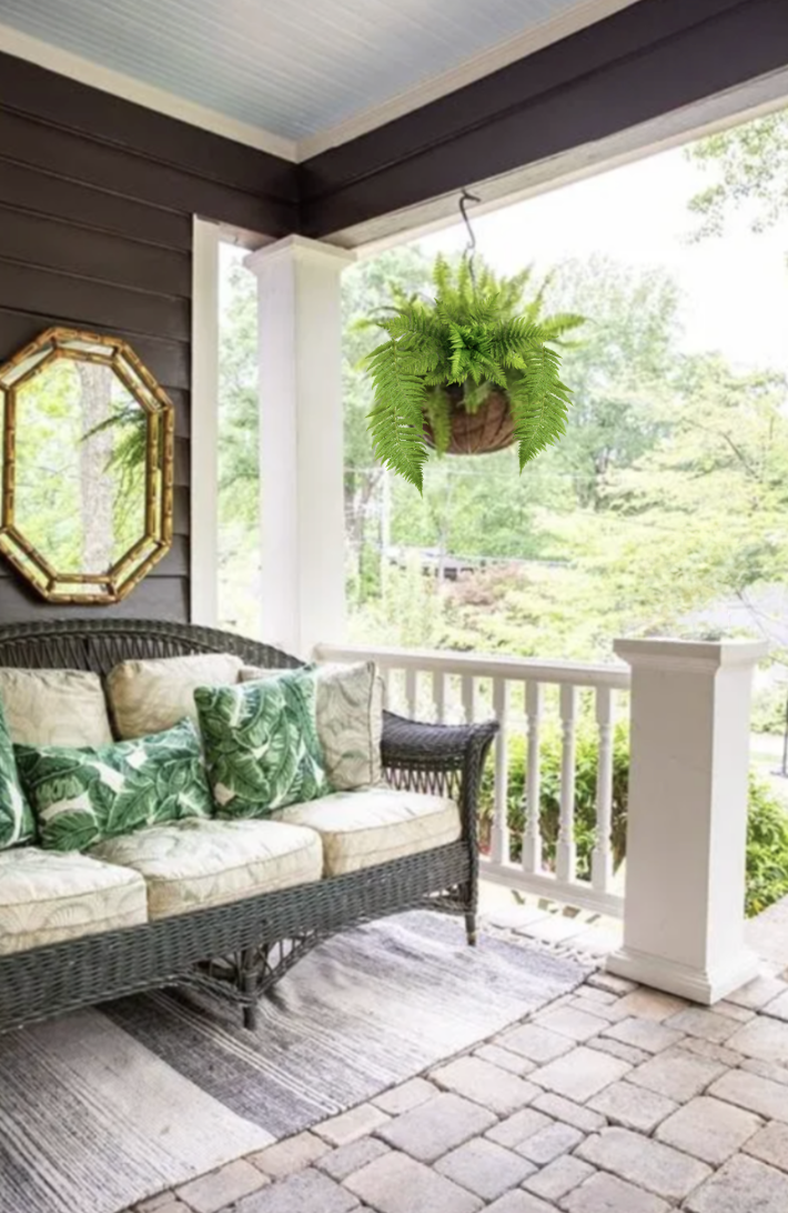 Porch with wicker sofa, tropical-pattern cushions, hanging fern plant, mirror, brick flooring, and railing with view of green trees outside.
