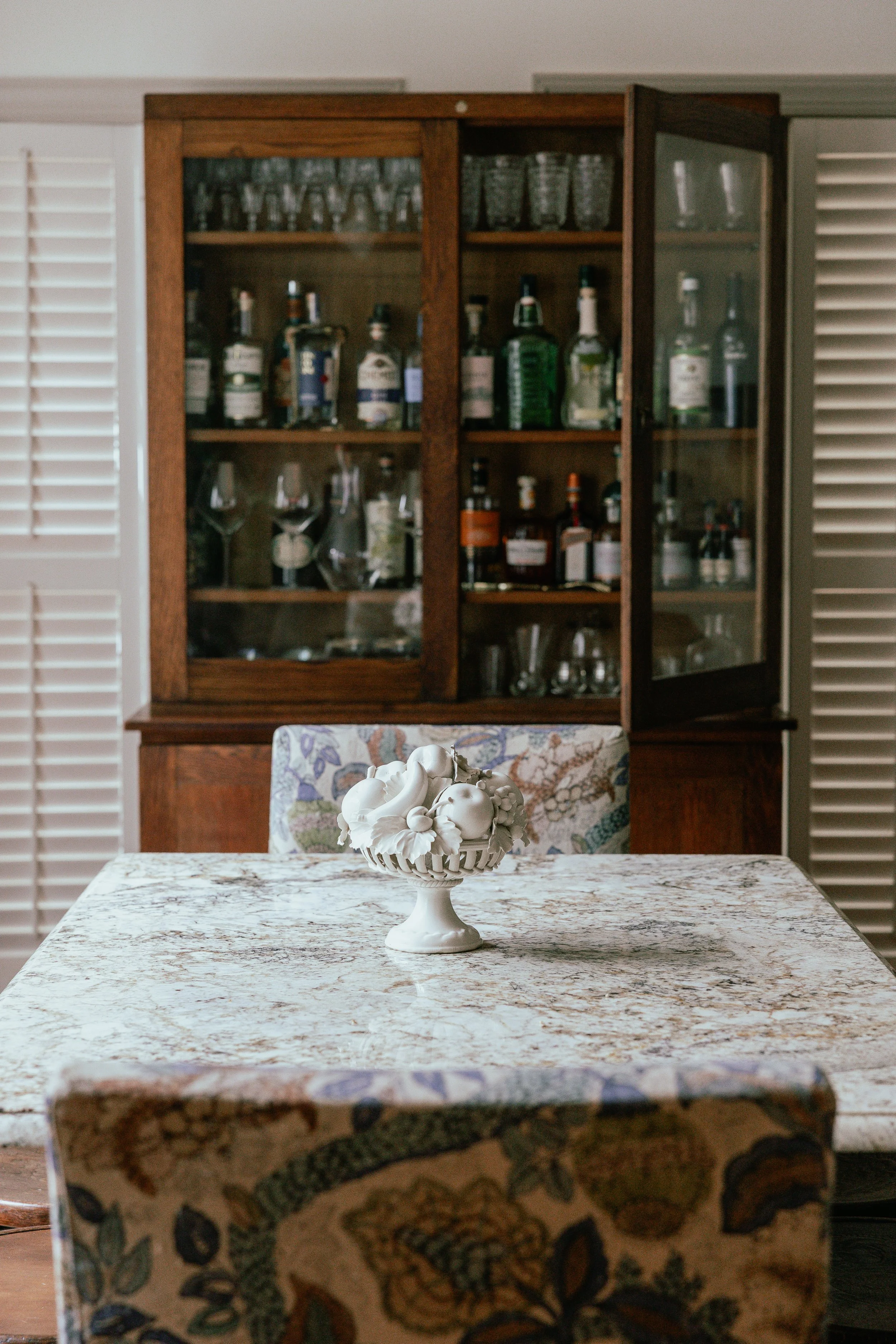 A marble dining table with a ceramic fruit bowl centerpiece, with a hardwood glass-door cabinet filled with liquor bottles and glassware in the background.