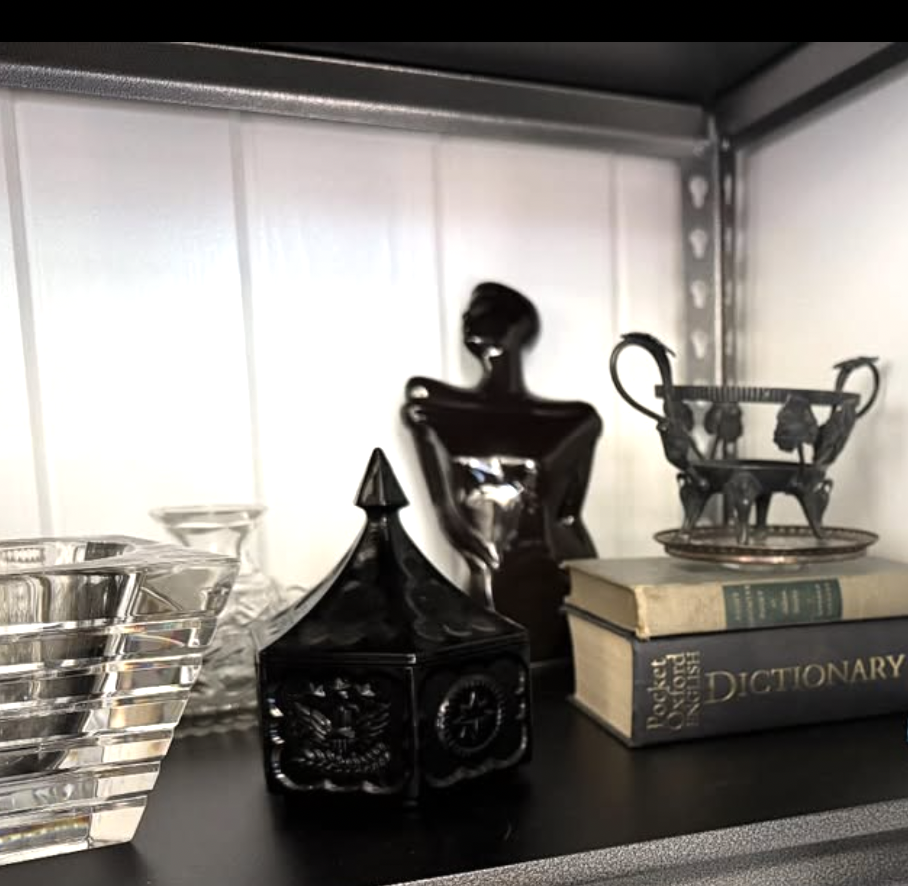 Decorative objects on a shelf, including a black sculpture of a woman, a black ornamental house with a pointed roof, a clear glass dish, a small ornate glass cup, an old hardcover dictionary titled 'Dictionary,' and a small glass dish stacked on top of the dictionary.