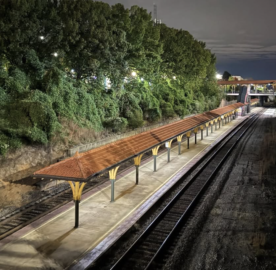 Empty train station platform at night, with a canopy roof and surrounding green trees. Visible railroad tracks run alongside the platform.