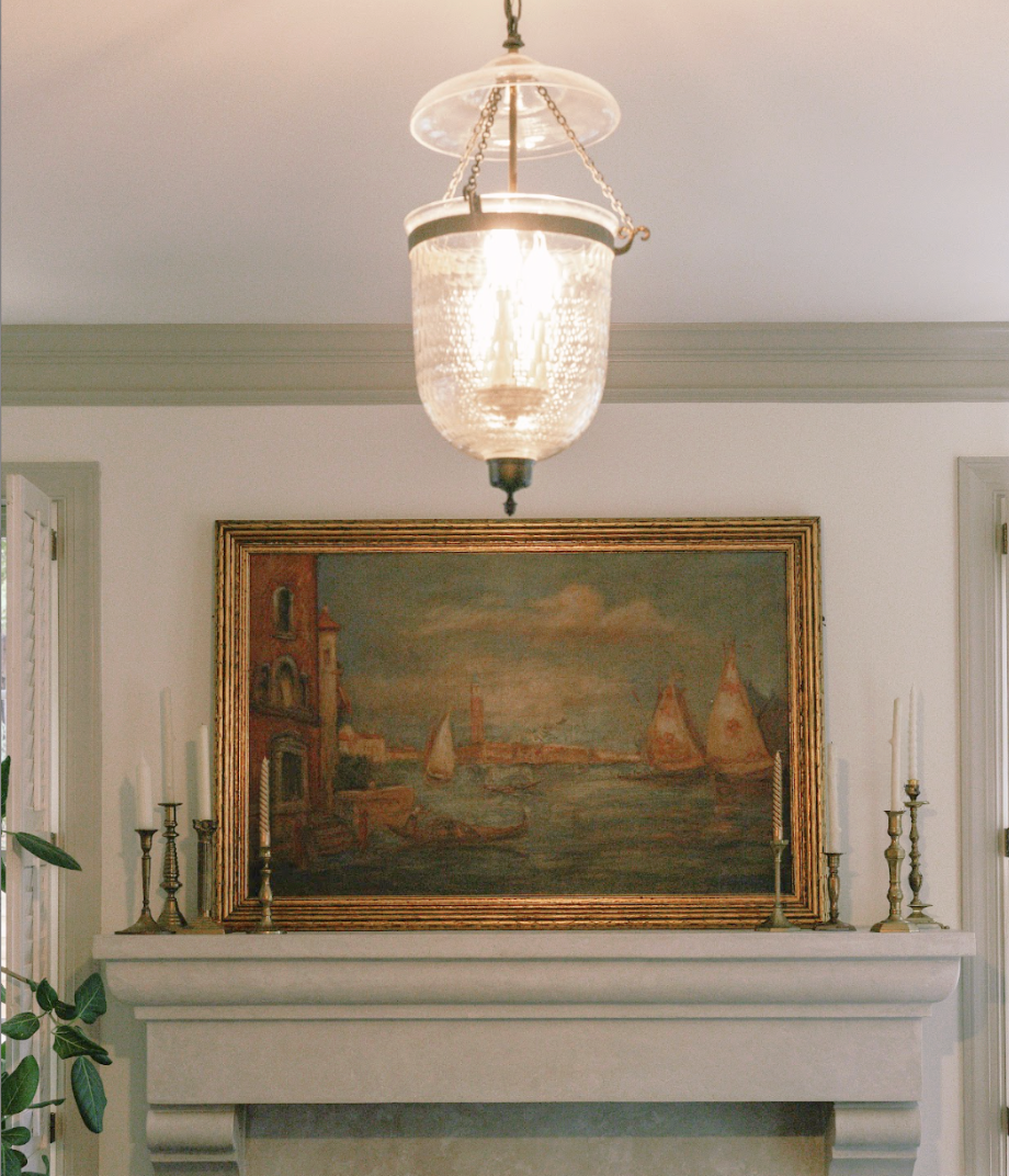 A vintage glass chandelier hanging from a ceiling, illuminating a decorative stone fireplace with an oil painting of a harbor scene, framed in gold, above it. The fireplace mantel is decorated with brass candlesticks, and a potted plant is partially visible on the left.