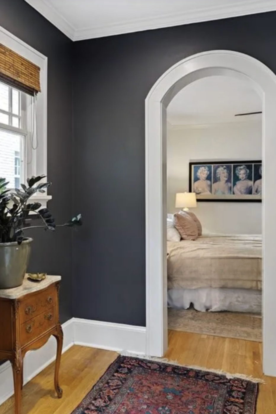 View of a corner of a room with dark gray walls, a window with bamboo blinds, and a small ornate wooden side table with a potted plant. Through an arched doorway, a bedroom with a bed, a table lamp, and a framed art piece of Marilyn Monroe is visible.