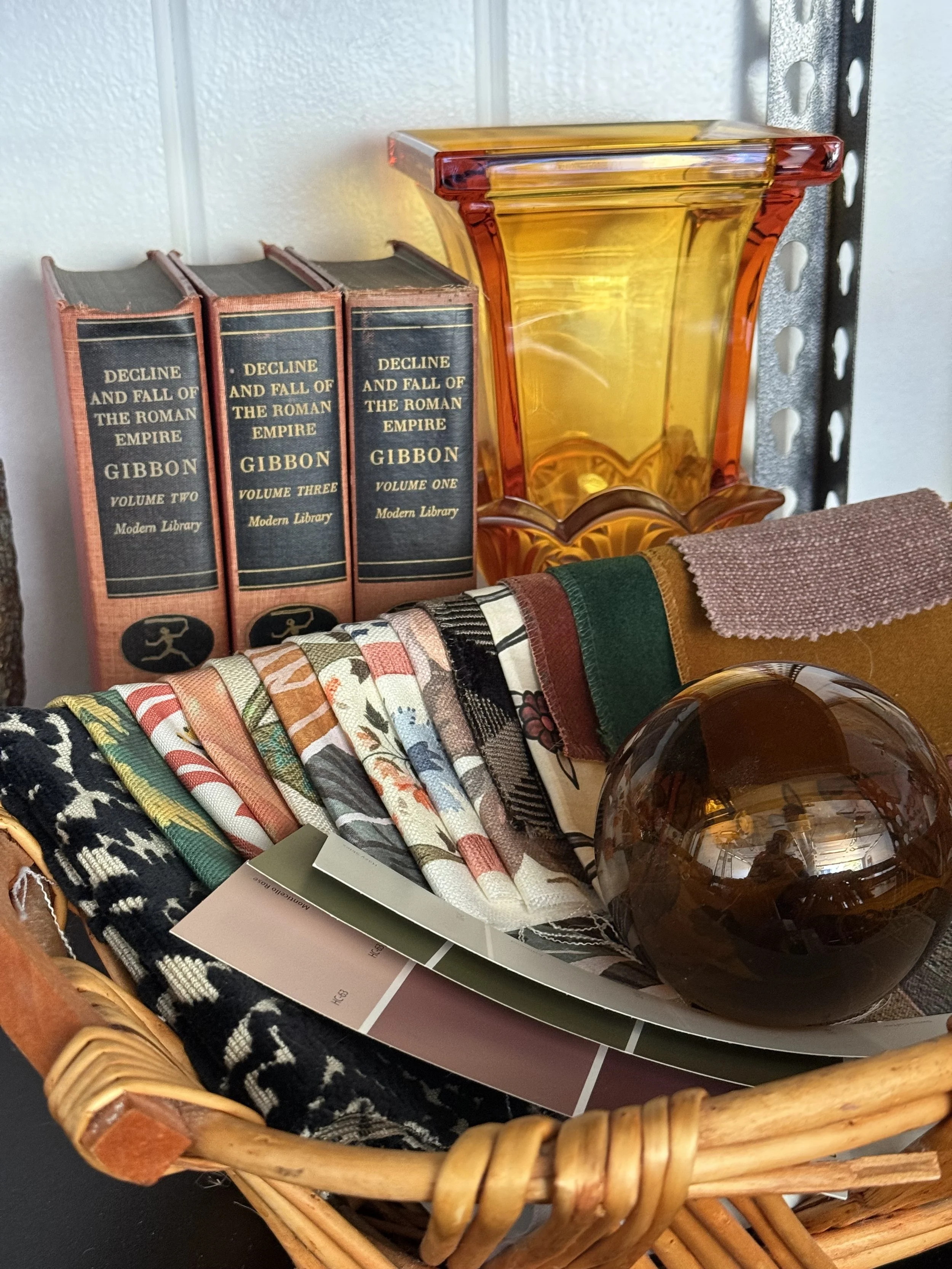 A wire basket filled with fabric swatches and color samples, a large brown glass paperweight, and sample color sheets. Behind the basket, a stack of three books titled 'Decline and Fall of the Roman Empire' by Gibbon, with volumes labeled one, two, and three, and a large amber-colored glass vase on a metal shelf.