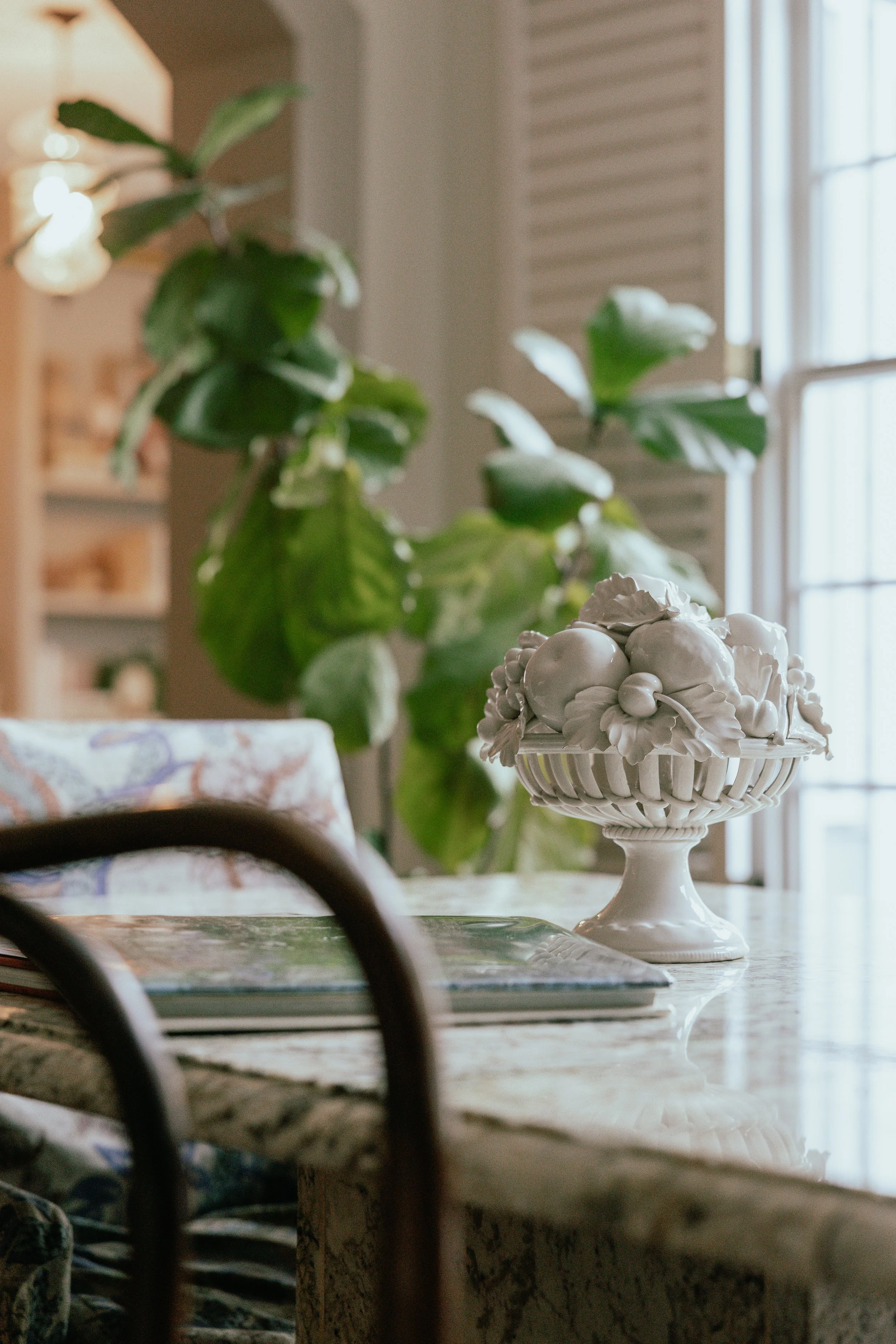 Indoor dining table with a decorative white ceramic fruit bowl filled with fruit, near a window with daylight, and a large green plant in the background.