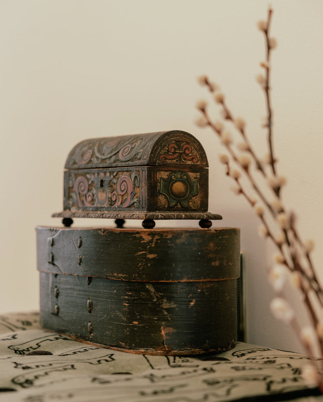 A vintage wooden jewelry box with intricate carvings and colorful painted patterns, placed on a distressed black box, with a branch of pussy willow flowers in the foreground.