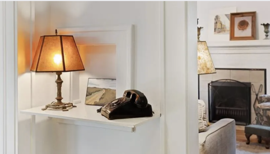 Living room with a white built-in shelf holding a table lamp and a vintage rotary phone, with a sofa, fireplace, and framed pictures in the background.