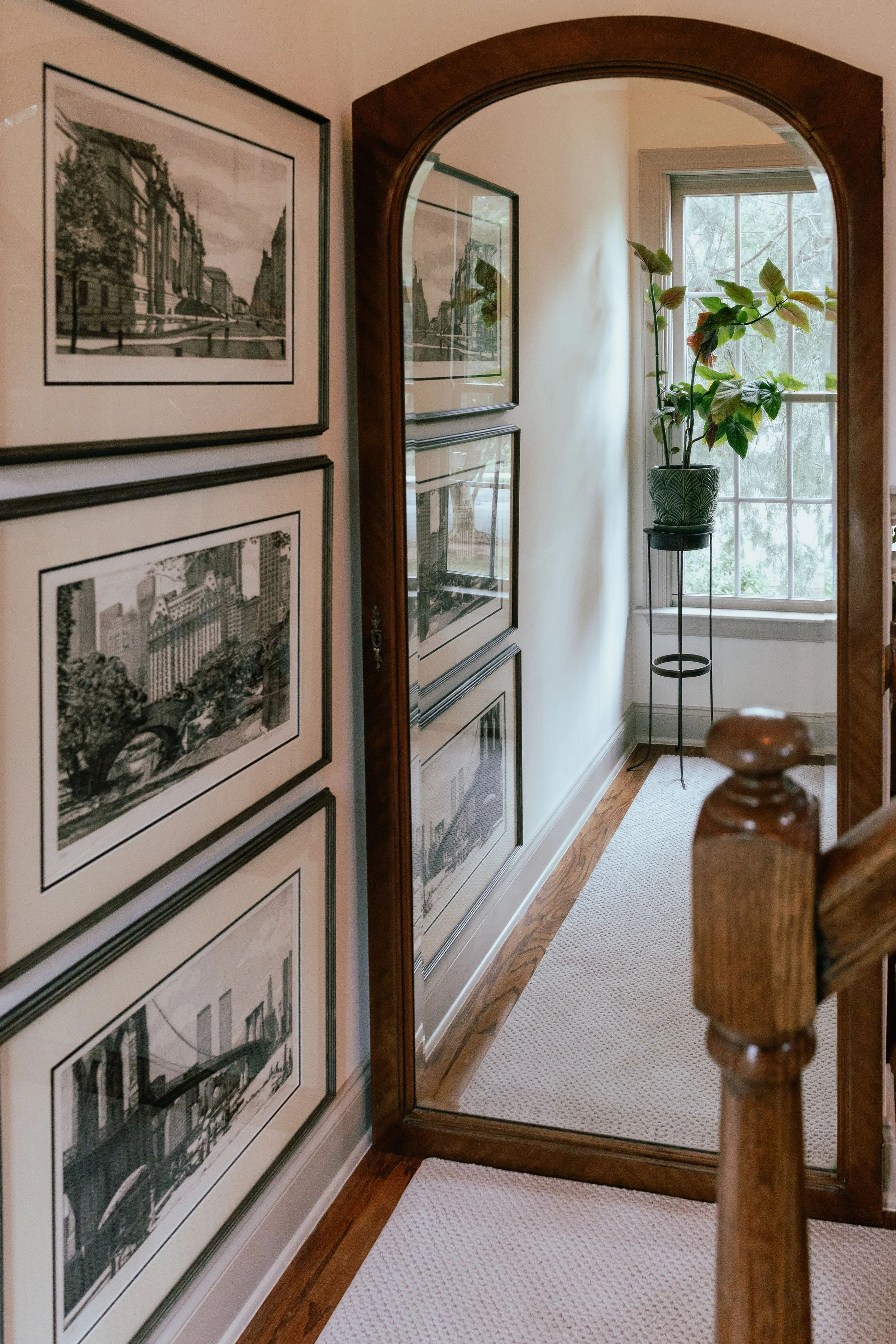 A hallway with a large wall-mounted mirror, artwork of cityscapes on the left wall, a plant in a decorative pot on a stand, a window letting in natural light, and a white runner rug on hardwood floors.
