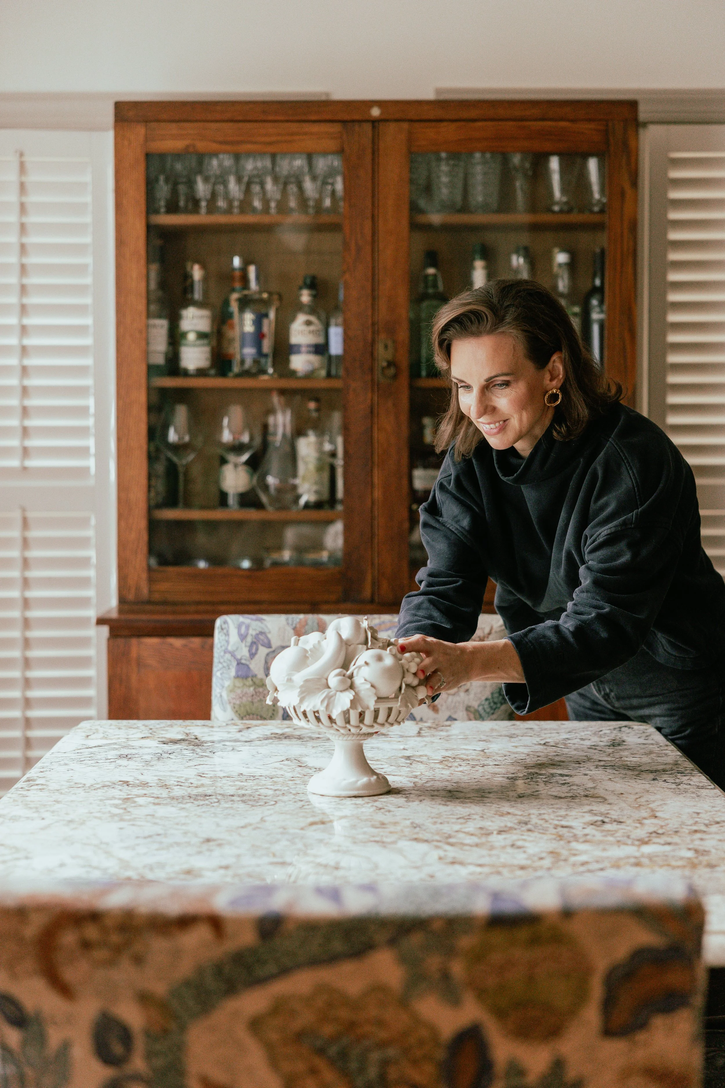 A woman in a black shirt arranging a white decorative bowl on a marble dining table in a dining room with a wooden cabinet filled with bottles and glasses in the background.