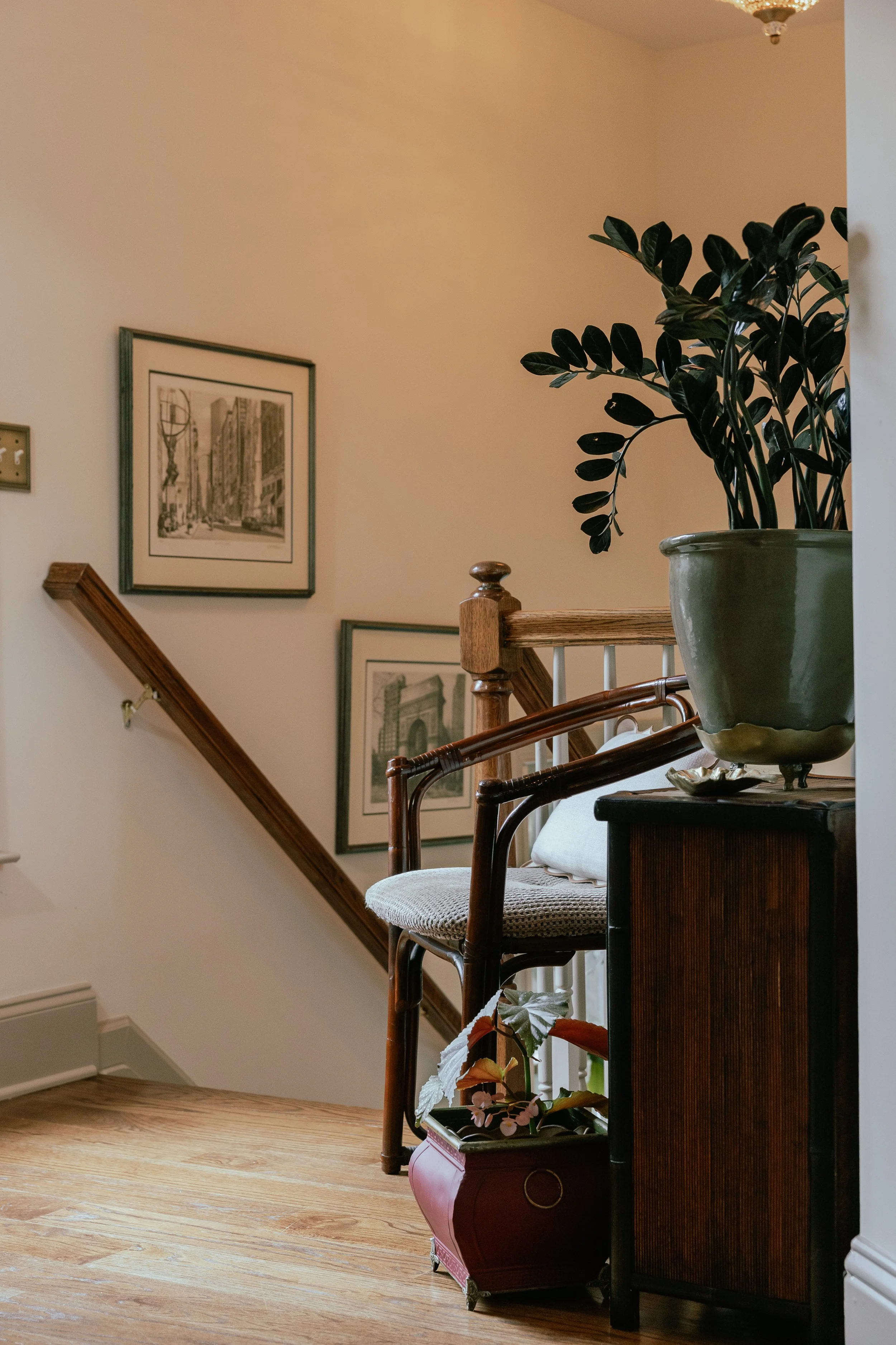 A cozy corner of a home with a wooden staircase, framed artwork on the wall, a chair with a cushion, and a large potted plant on a cabinet.