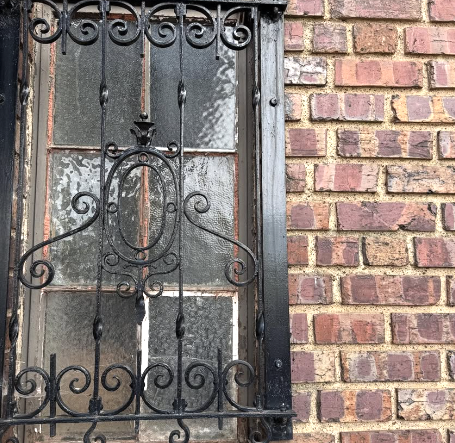 A narrow window with frosted glass panes, surrounded by a black wrought iron security grille with decorative scrollwork, mounted on a brick wall.