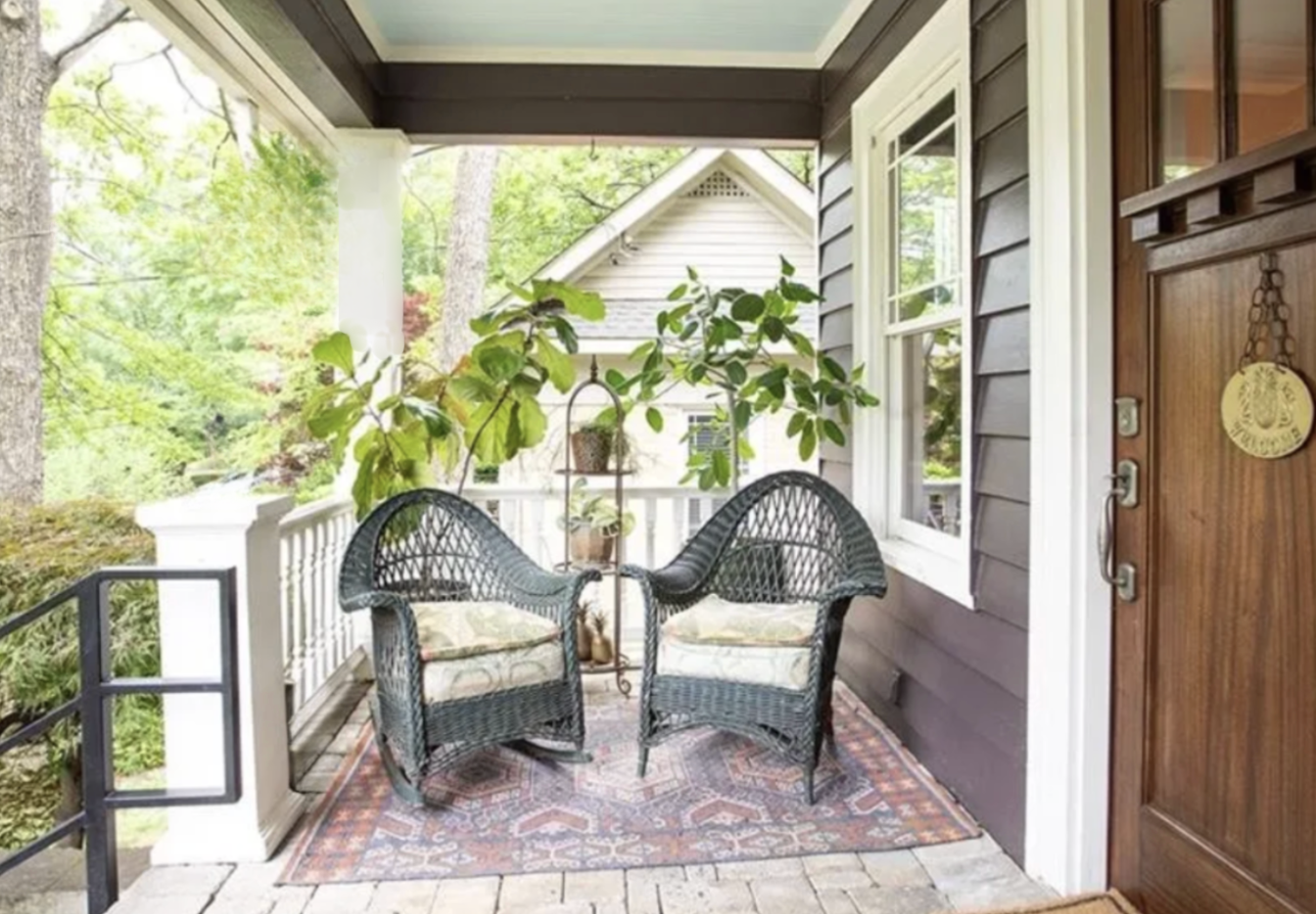 A cozy front porch with two black wicker chairs, a small patterned rug, and a large green potted plant. The porch has a white railing and is attached to a house with gray siding and a wooden front door. Trees and greenery are visible in the background.