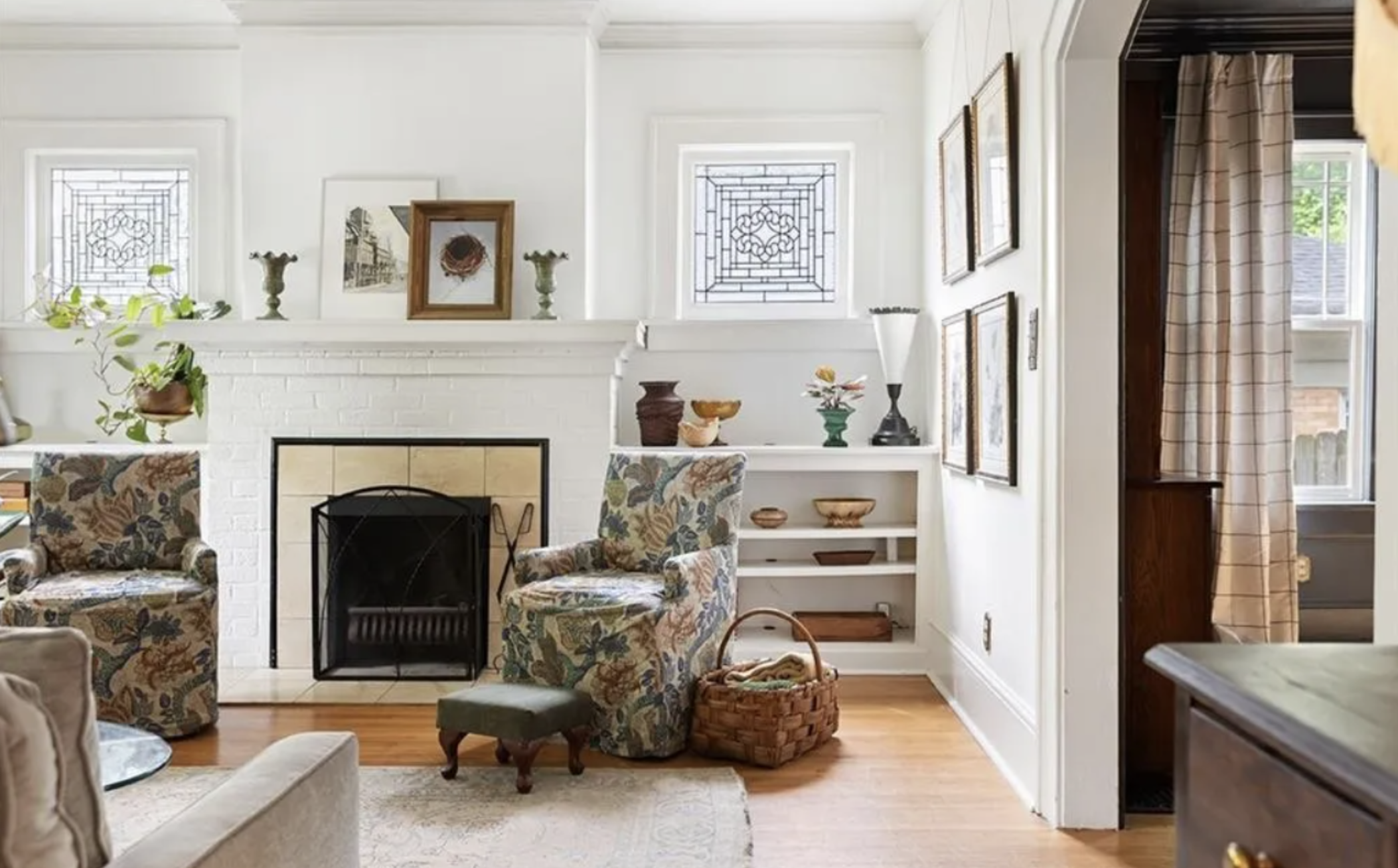 A cozy living room with a white brick fireplace, patterned armchairs, and various decorative items on the mantel and built-in shelves. There is a wicker basket on the hardwood floor, beside an ottoman, and sunlight coming through a nearby window with checkered curtains.