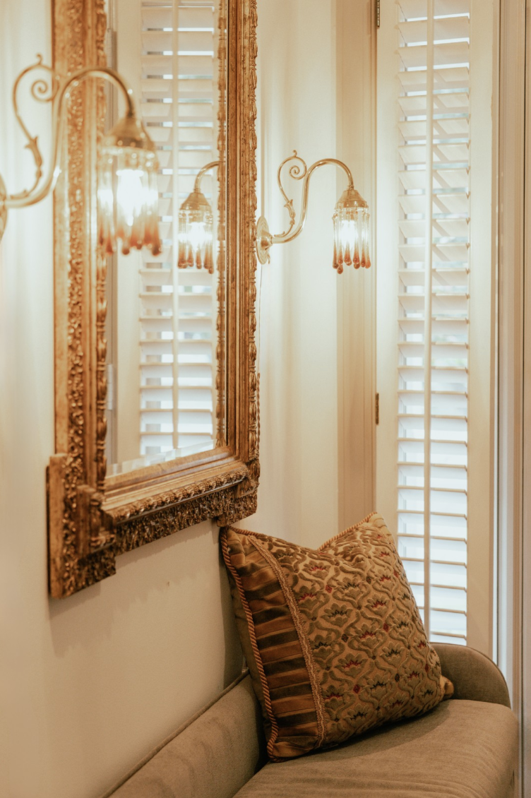 A cozy corner with a beige sofa, decorated with a patterned throw pillow, next to a wall-mounted vintage gold ornate mirror and a wall sconce with a decorative glass shade. White plantation shutters cover a window in the background.