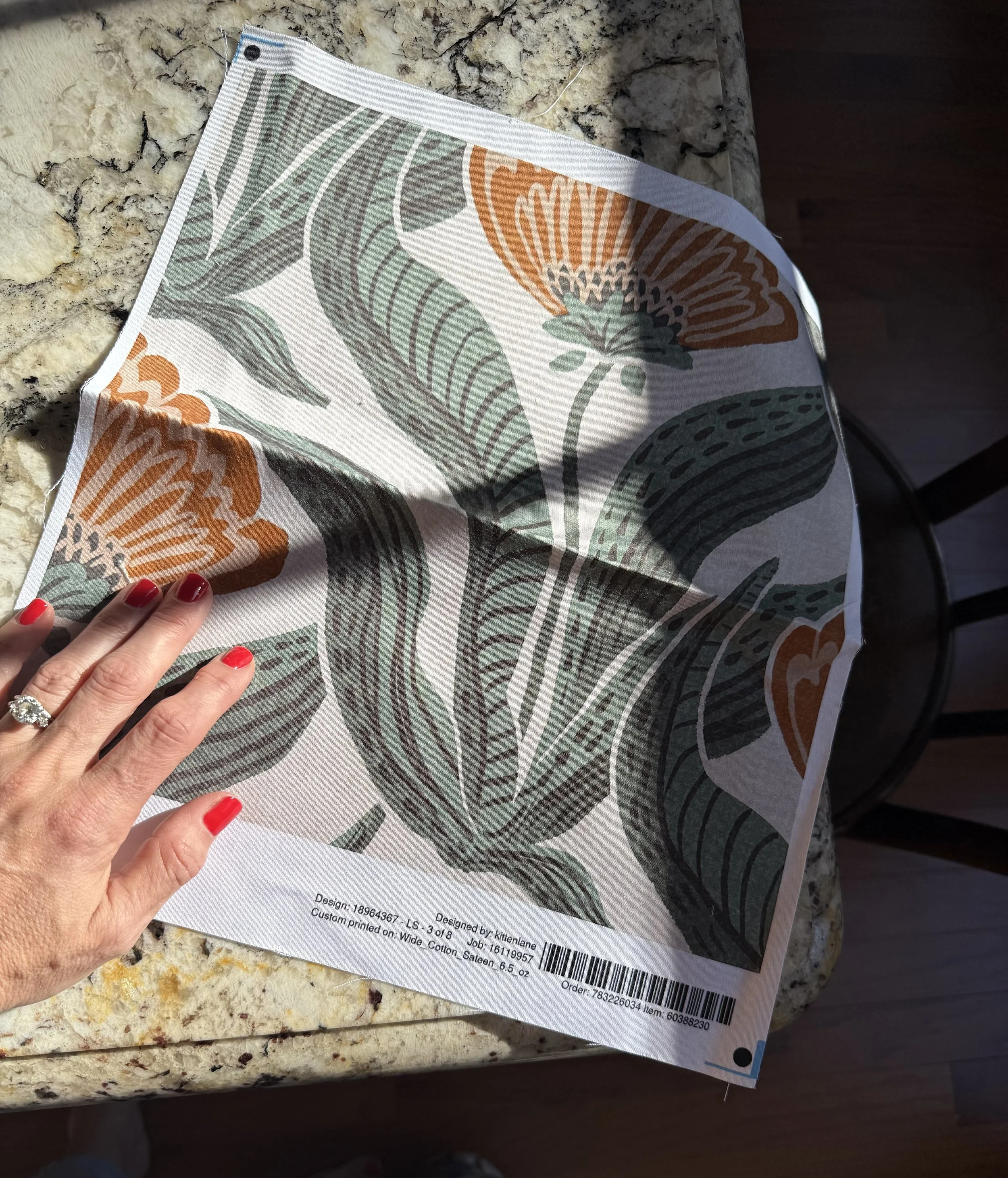 A person with red painted nails and a ring on their finger holding a fabric sample with a floral and leaf pattern in green, orange, and white, placed on a granite countertop.