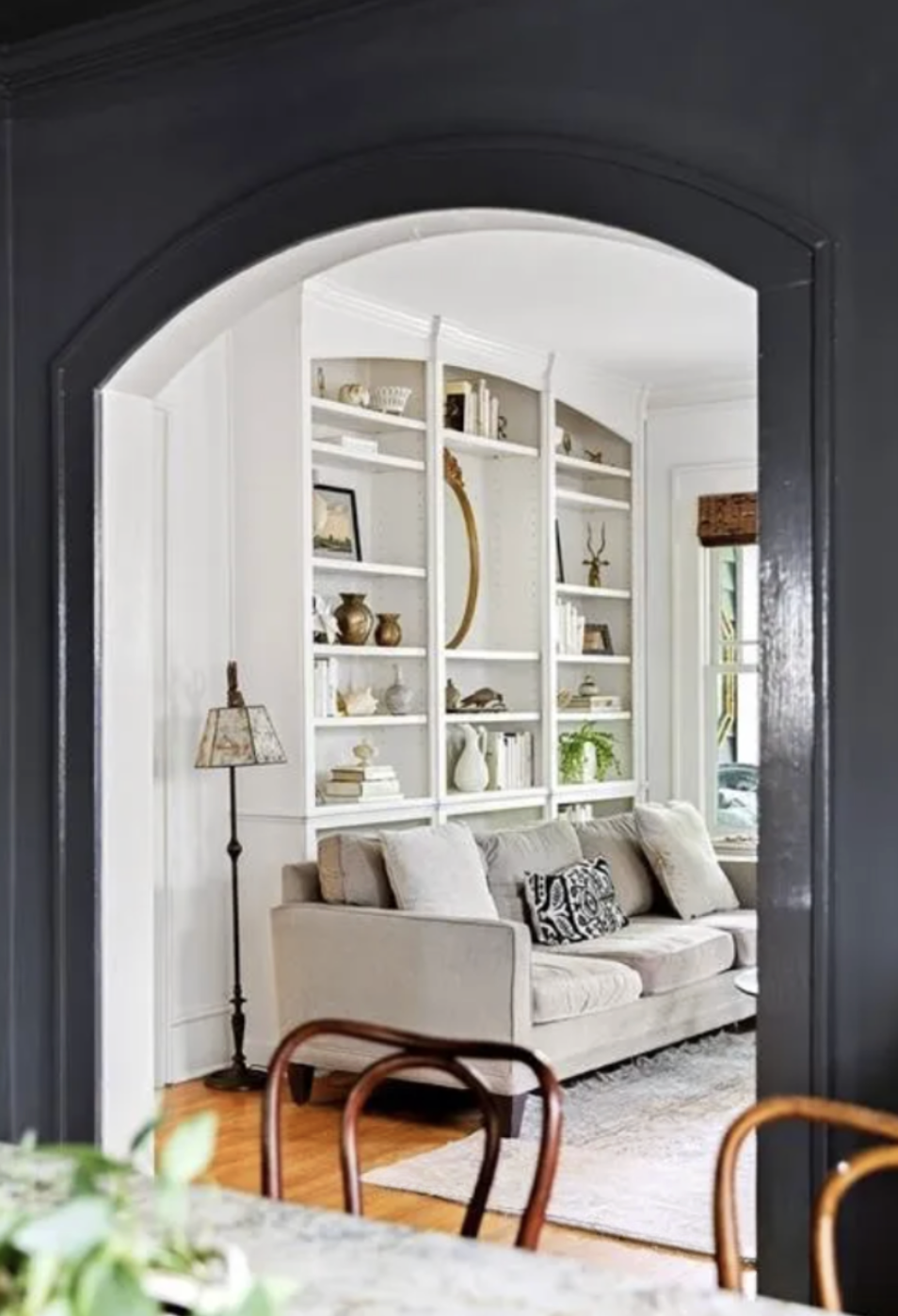Living room seen through an arched doorway, featuring a beige sofa with patterned and solid pillows, a white built-in bookshelf with decor and books, a window with a bamboo shade, and a floor lamp with a patterned shade.