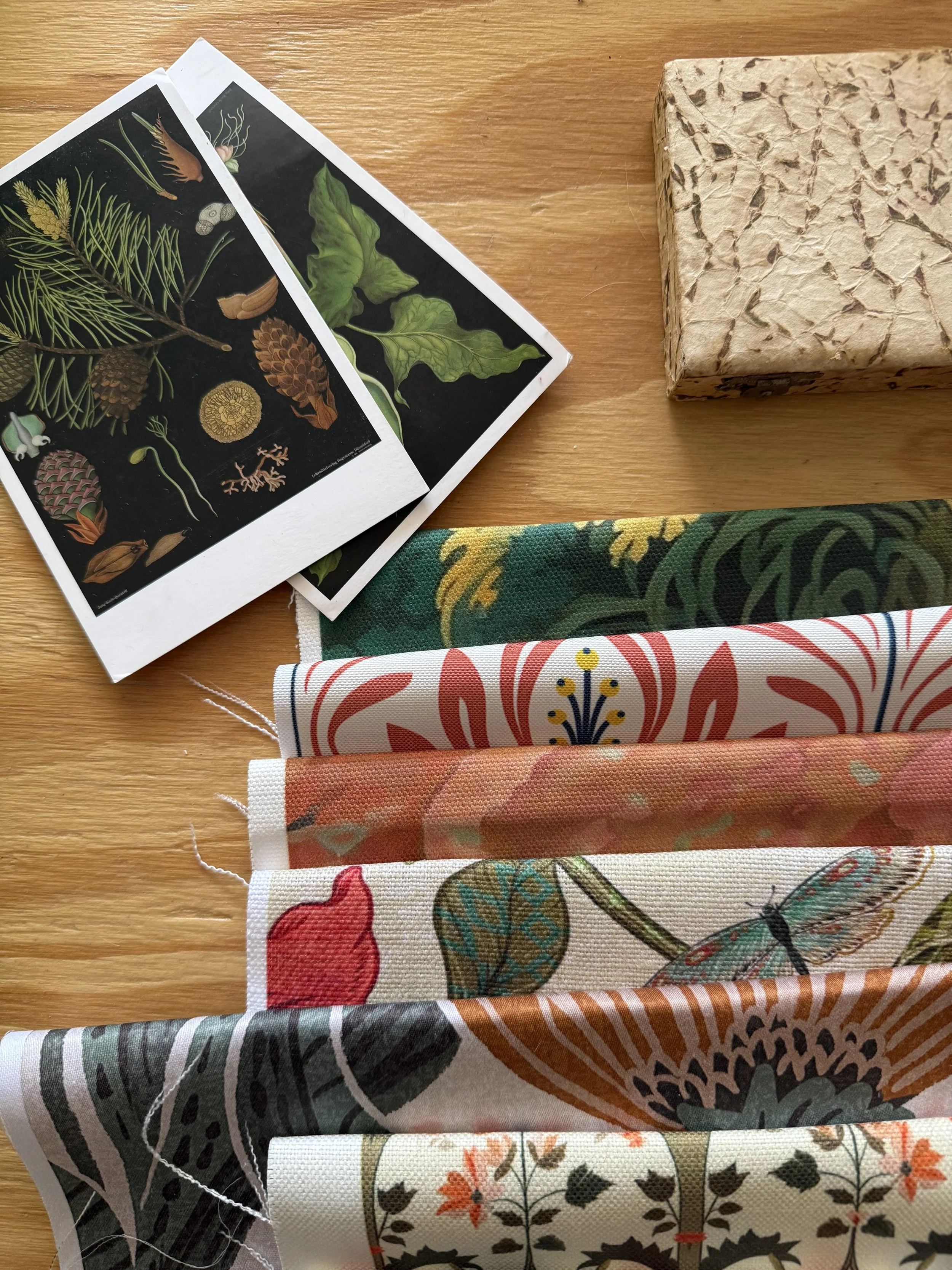 A wooden table holding botanical illustrations, a stone-patterned box, and five colorful fabric swatches with floral and insect designs.