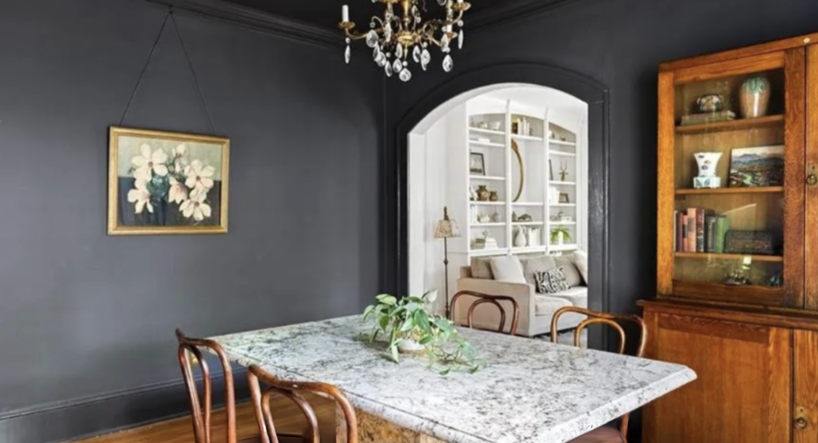 Dining room with dark gray wall, chandelier, a framed floral painting, a marble-topped dining table with wooden chairs, and a plant centerpiece. An open doorway reveals a living room with a beige sofa, a white bookshelf, and a lamp.