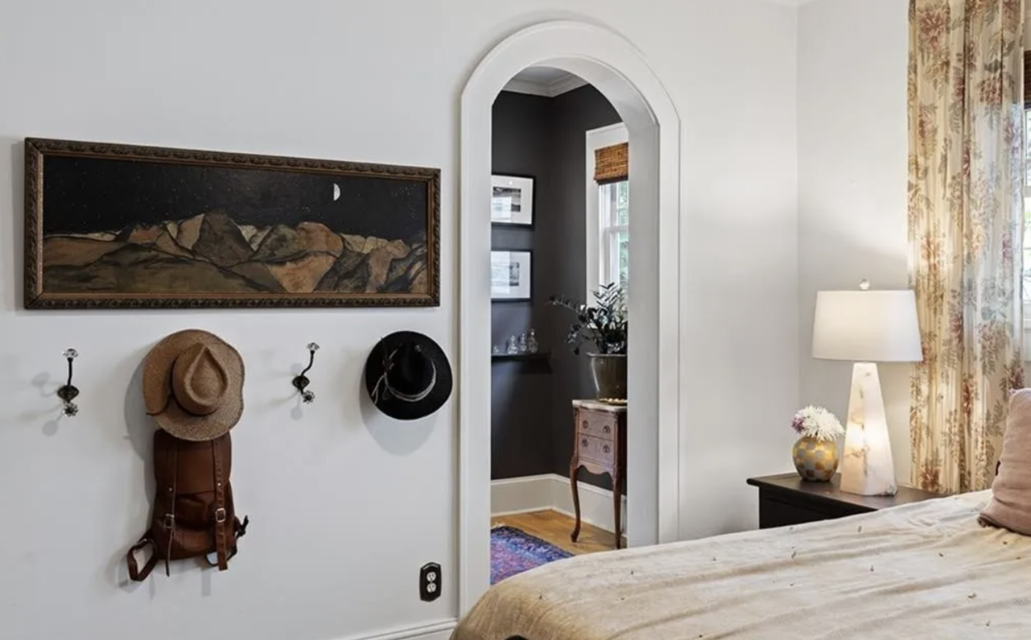 A bedroom area with white walls, a bed with beige bedding, a nightstand with a white lamp and a decorative vase with white flowers, a framed landscape painting, and a wall hook with a straw hat, a black hat, and a brown backpack. There is an arched doorway leading to a small room with dark walls, a window with a bamboo shade, a pink side table, and a plant.
