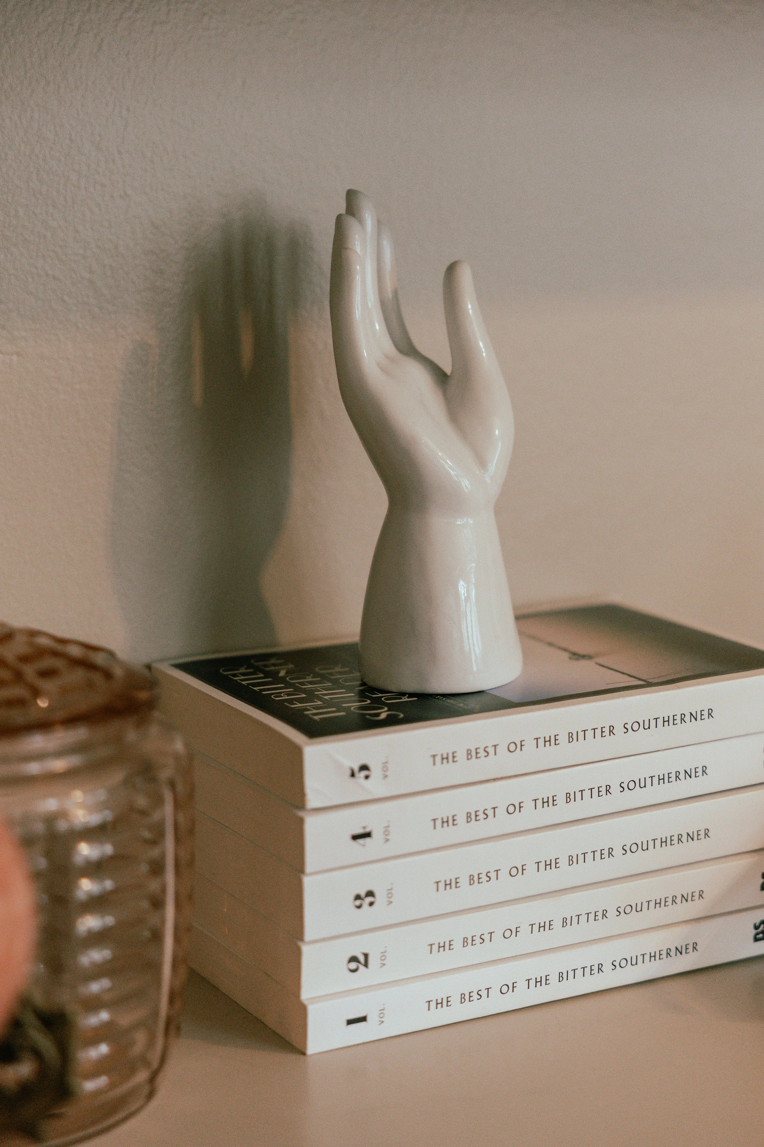 A white ceramic hand sculpture resting on top of a stack of five books titled 'The Best of the Bitter Southerner,' with a glass jar to the left.