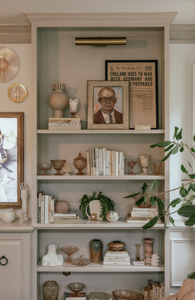 A white bookshelf filled with vintage glassware, books, and decorative pottery. The top shelf features framed artwork and a newspaper about World War I, with a narrow wall-mounted light fixture above. There are plants on the right side of the bookshelf and a partial view of a framed painting on the left.