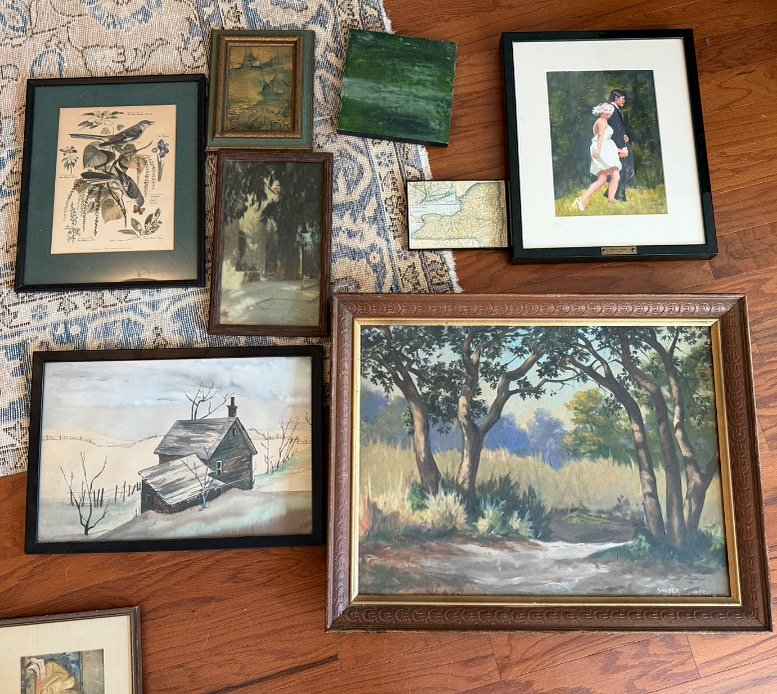 A collection of framed artwork and photographs laid out on a wooden floor. The pieces include landscape paintings, a vintage botanical print, and a photograph of a girl in a dress walking outdoors.
