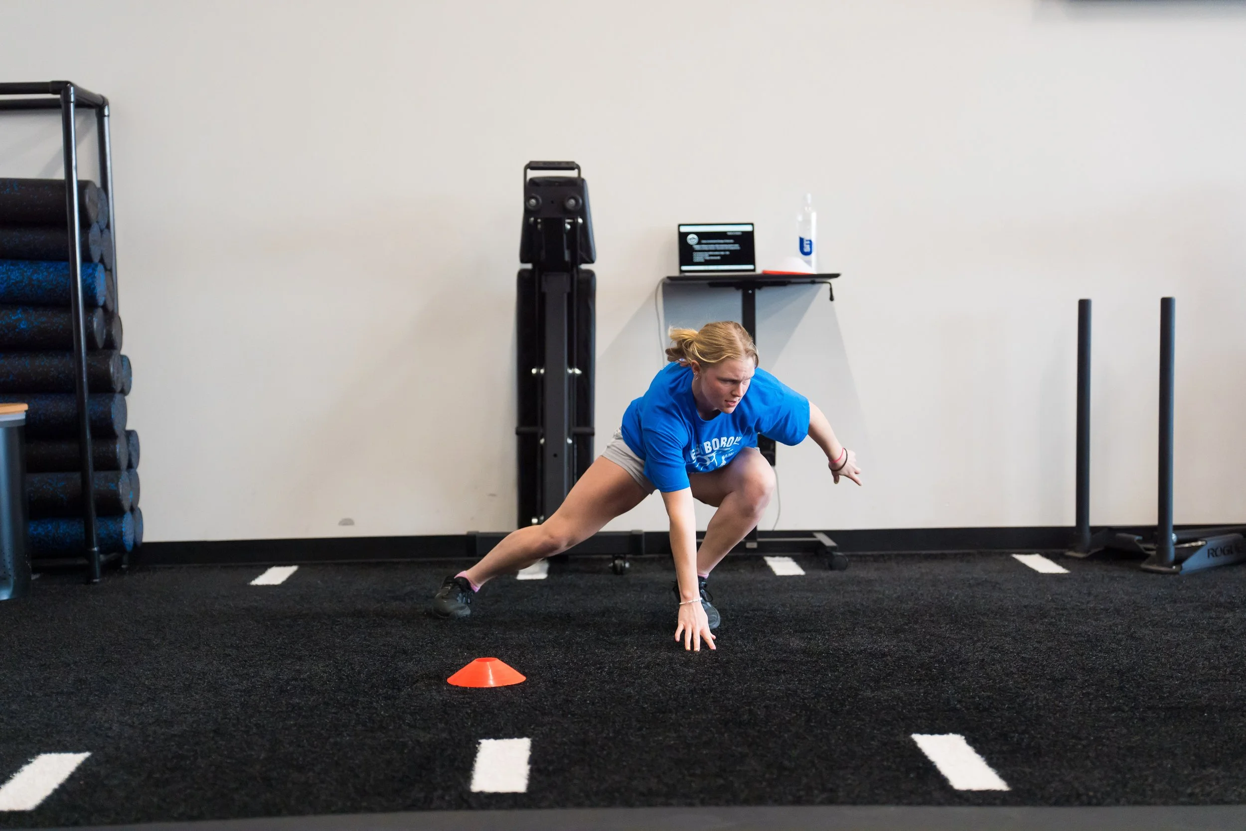 A woman performing a lateral movement exercise in a gym, crouched low with one arm reaching forward and the other down, near orange training cones on black rubber flooring.
