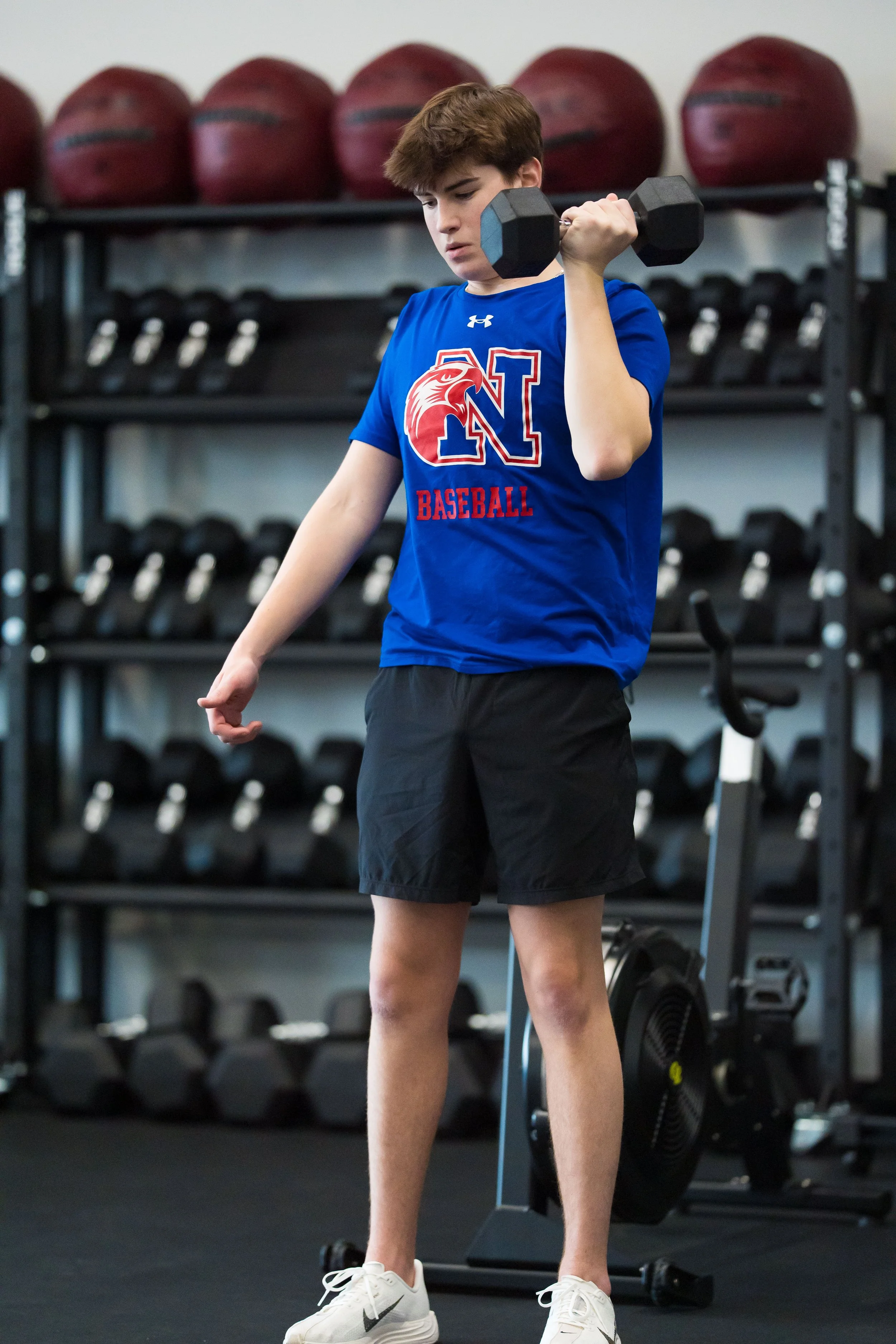 A young male athlete in a blue 'North' baseball shirt and black shorts lifting a dumbbell in a gym with kettlebells and medicine balls in the background.