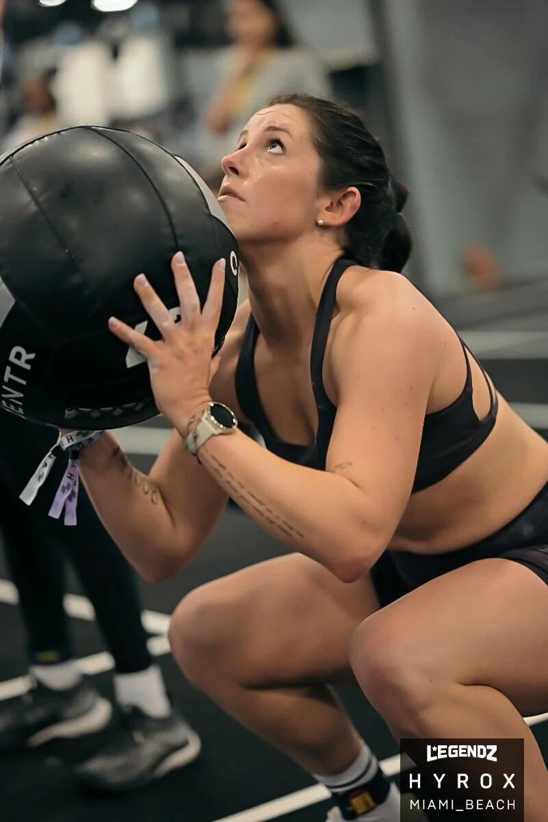 A woman in workout attire is mid-squat, holding a medicine ball close to her chest, doing a HYROX race