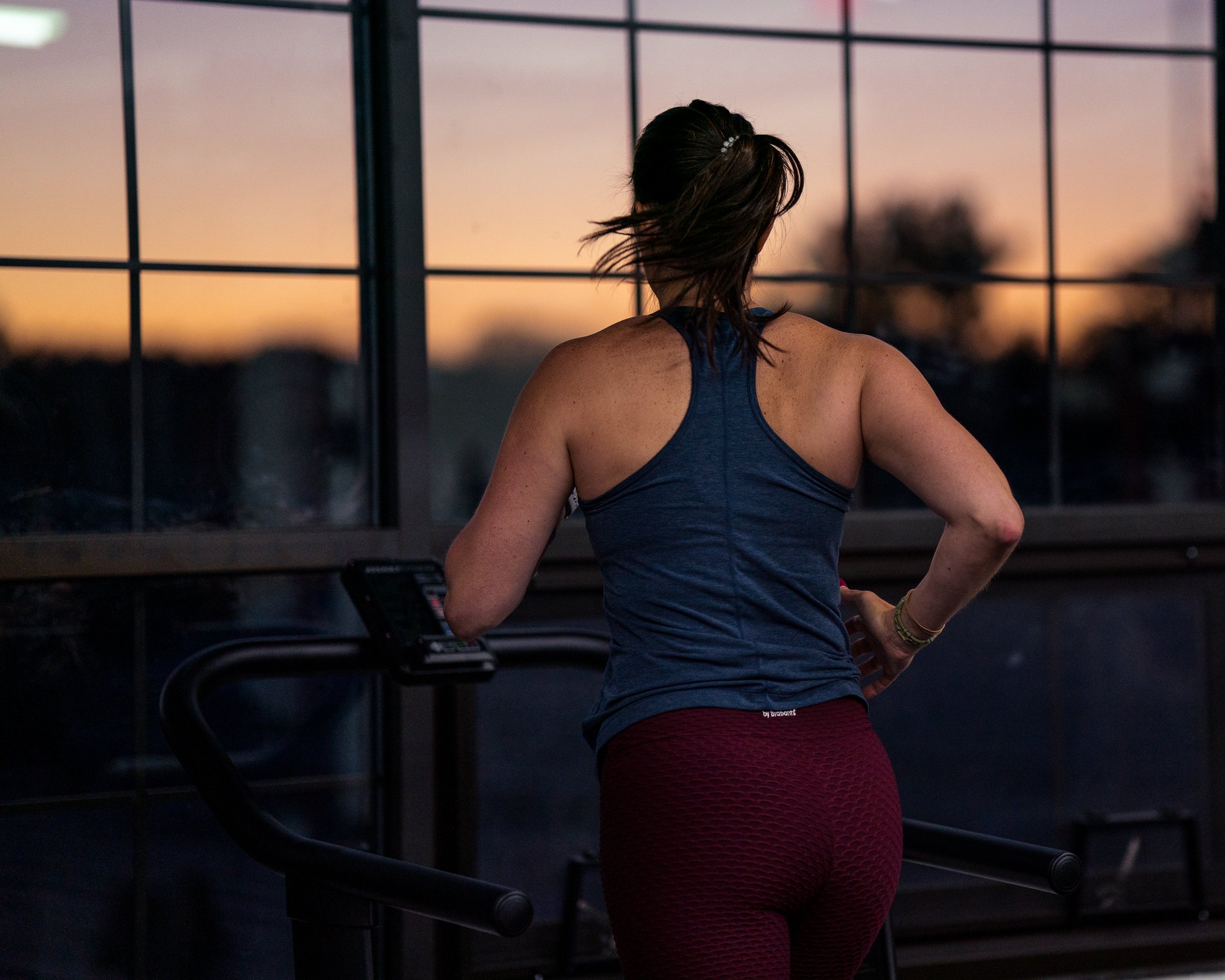 A woman in workout clothes standing on an indoor treadmill at sunset, looking out the window.