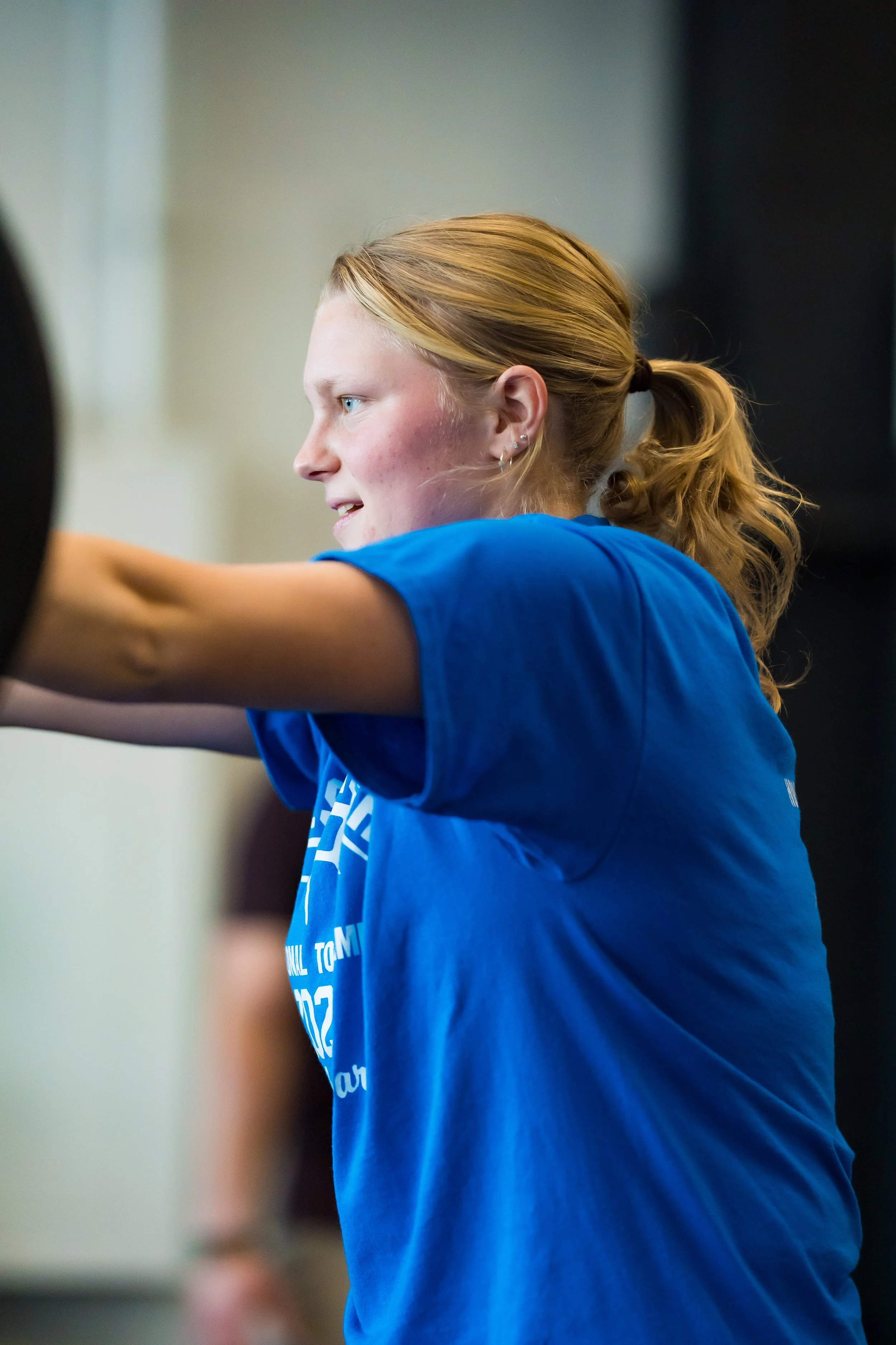 A young woman with red hair tied in a ponytail, wearing a blue t-shirt, appears to be exercising or lifting weights in a gym.